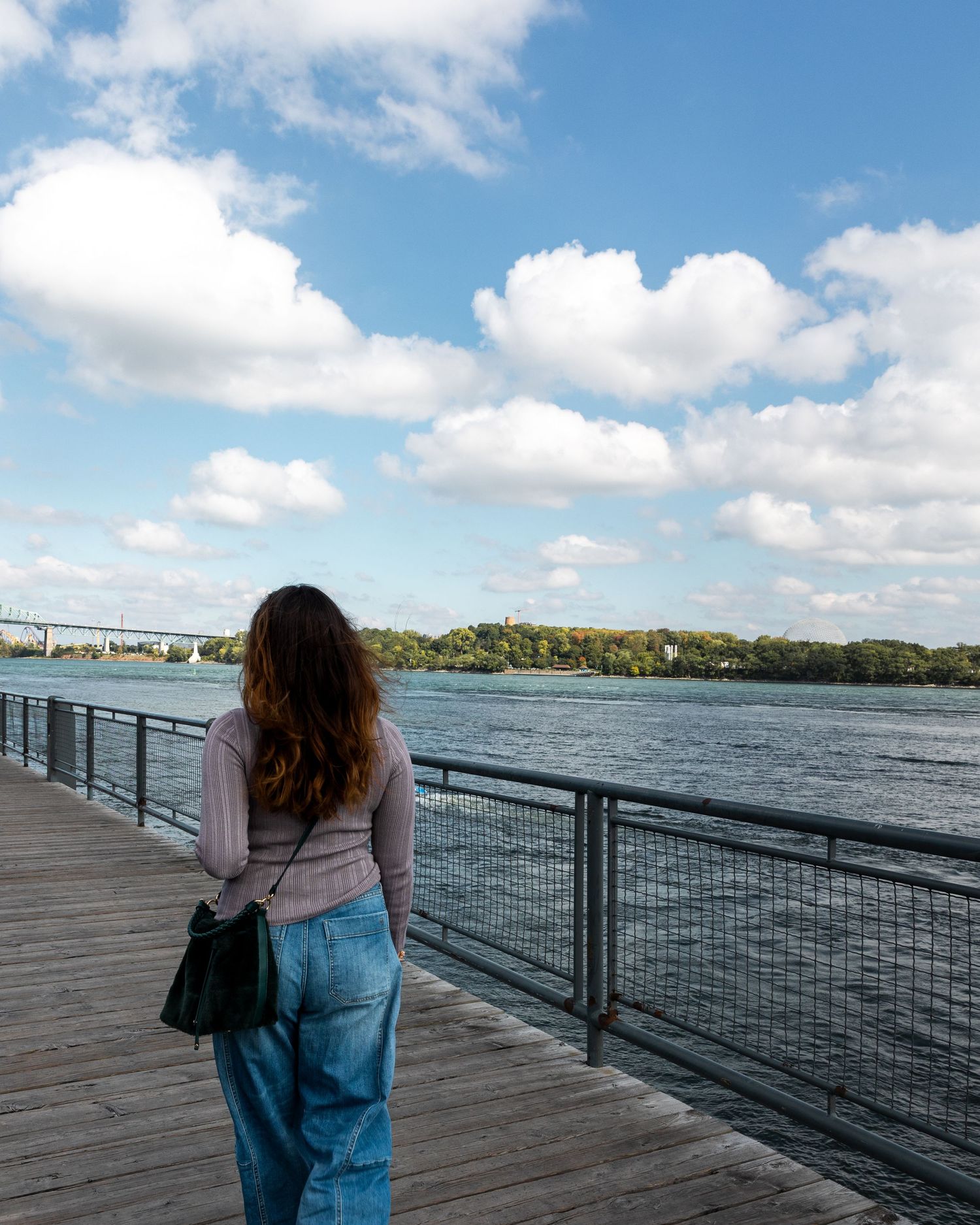 A person standing on a wooden deck overlooking a river and bridge on a sunny day with fluffy clouds in the sky.