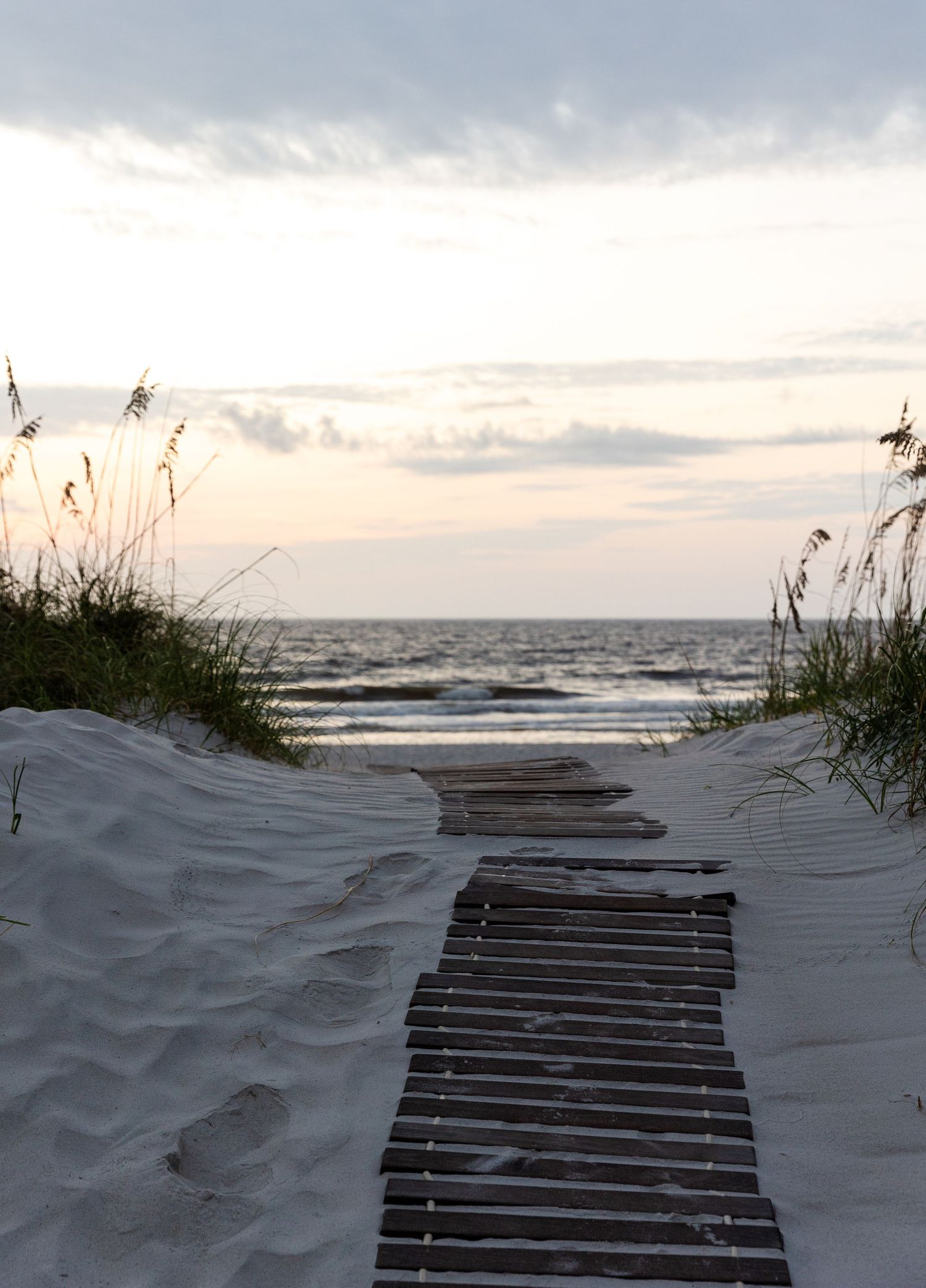 A wooden boardwalk path through white sand dunes leads to a beach at sunset with sea oats framing the view.