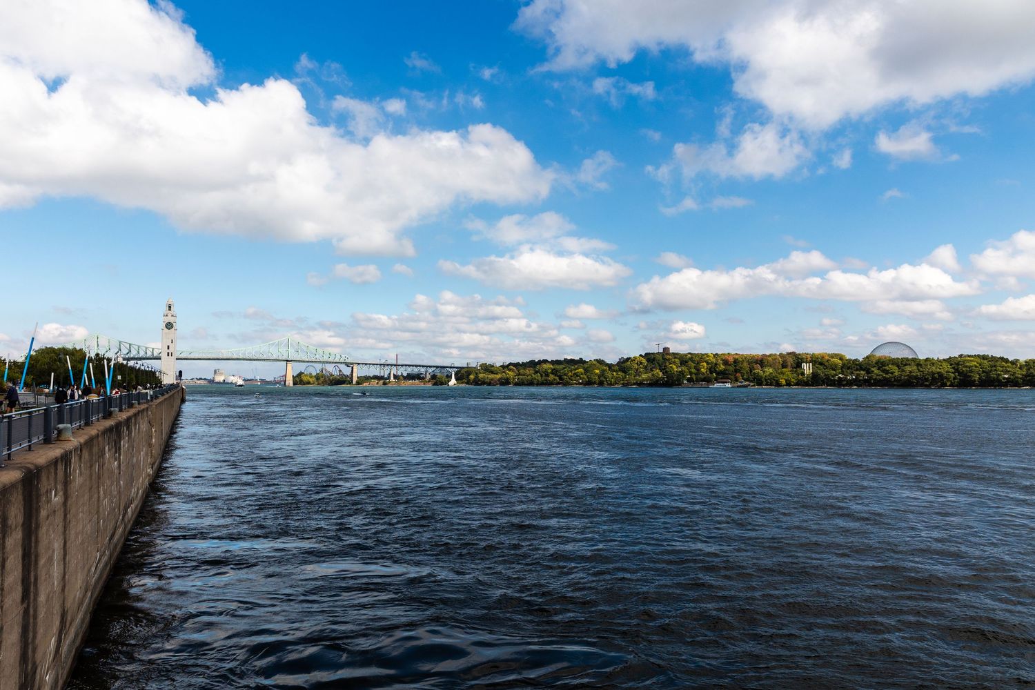 Large river with a bridge and walkway under blue skies with puffy white clouds in Montreal.