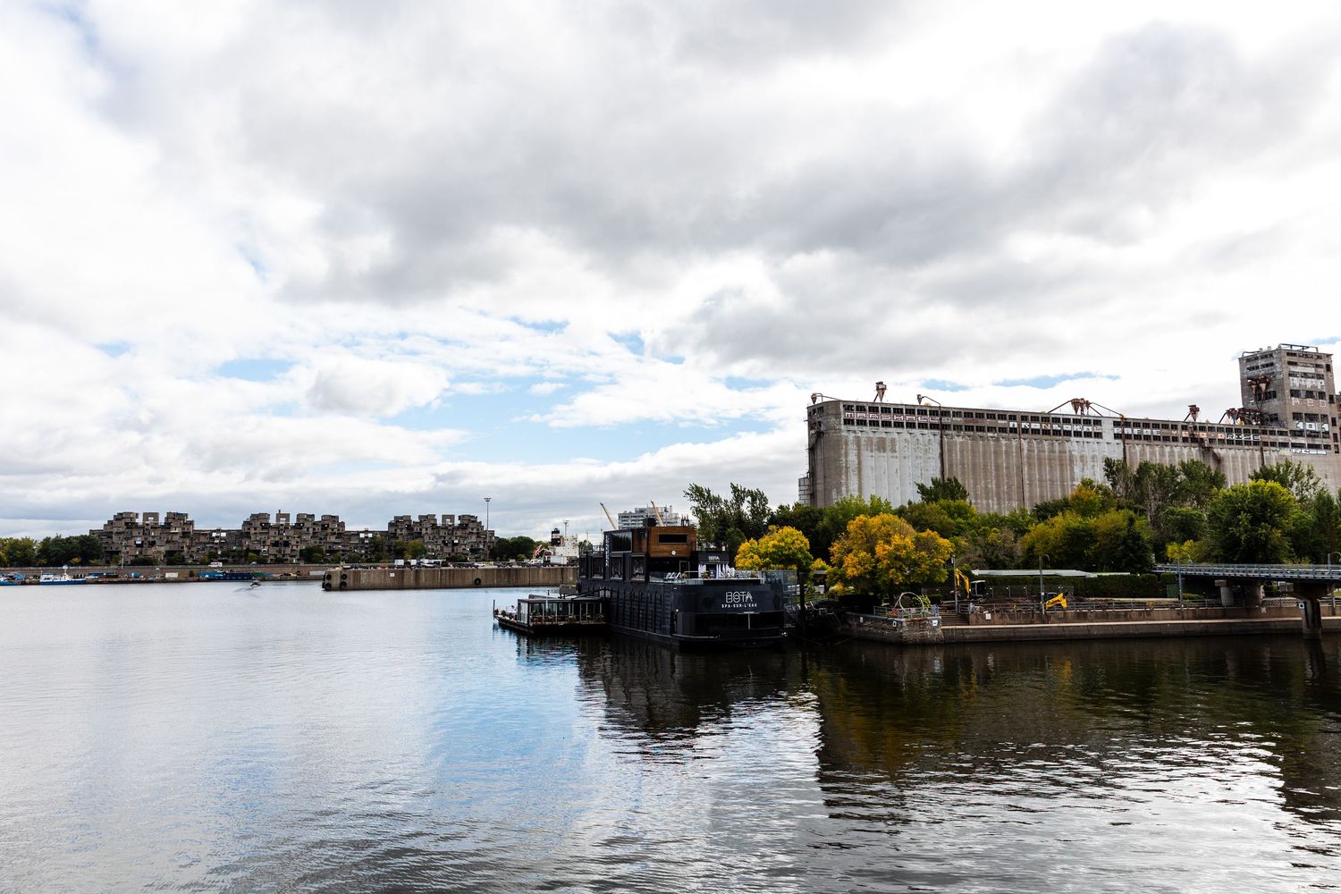 Industrial silos and buildings along calm waterfront with moored boats under cloudy sky in port setting.