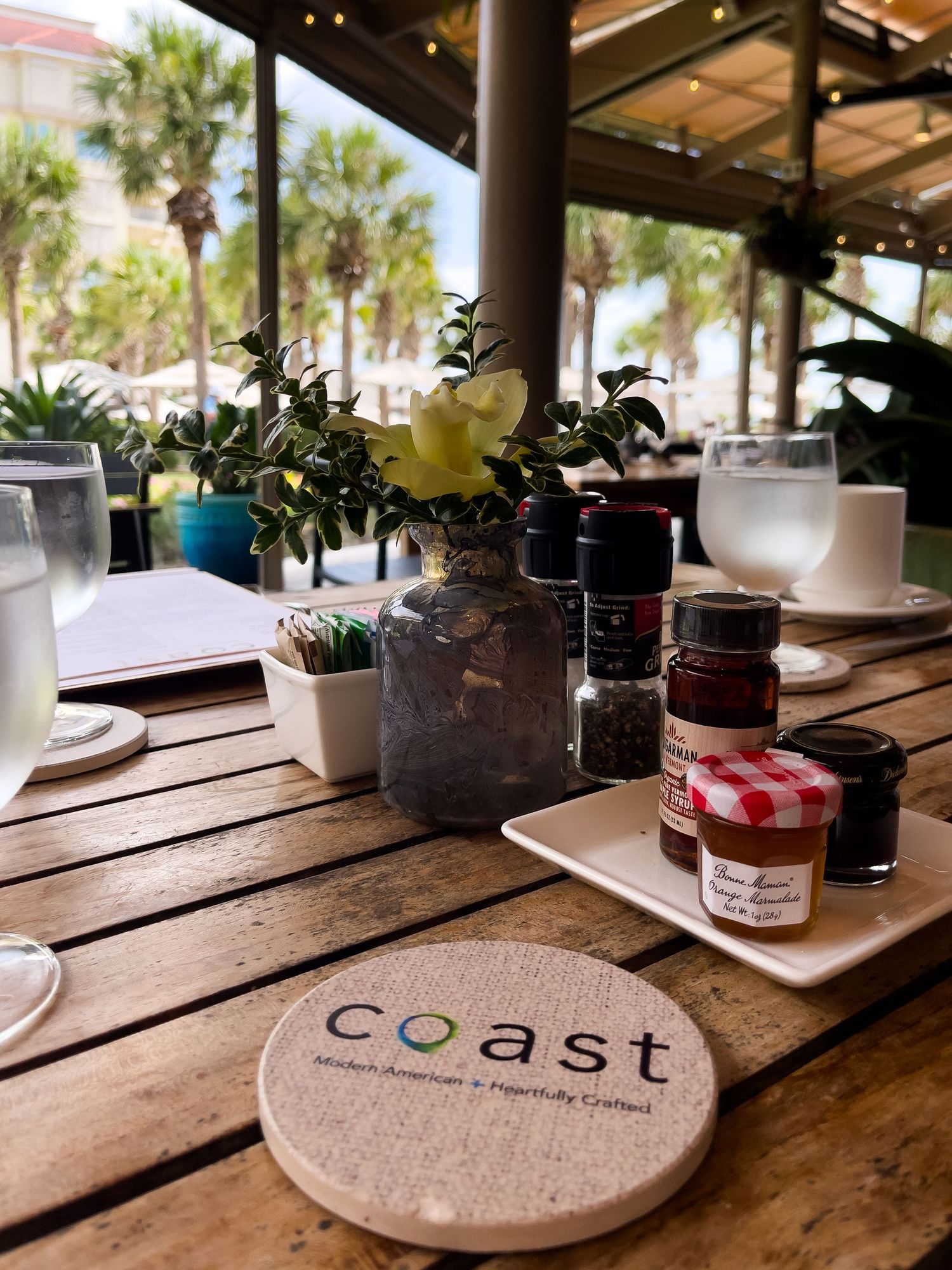 Restaurant table setting at Coast with elegant wooden decor, small flower arrangement, and jars of preserves by beachside palm trees.