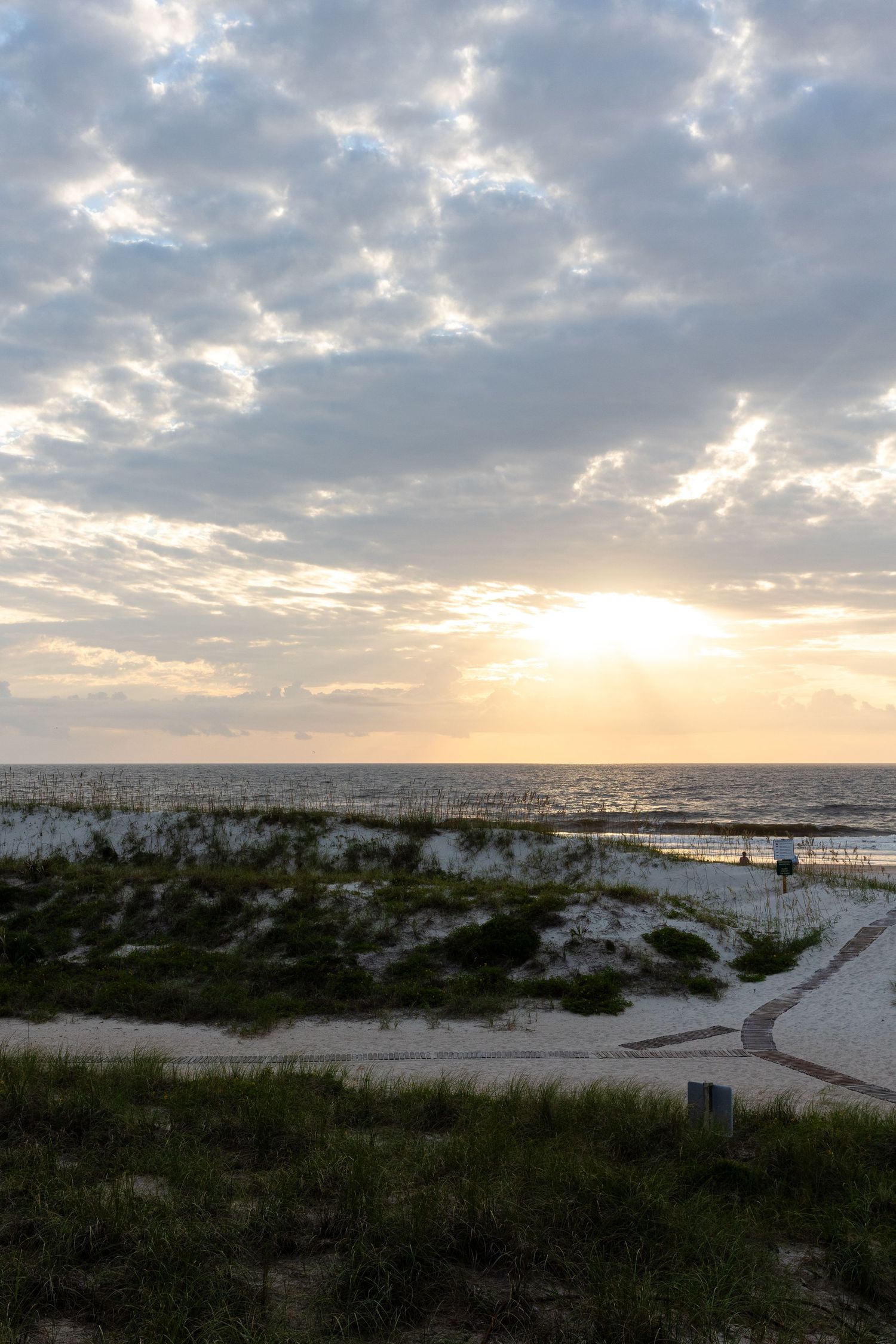 Scenic beach sunset with clouds casting rays over dunes and coastal vegetation along the shoreline.
