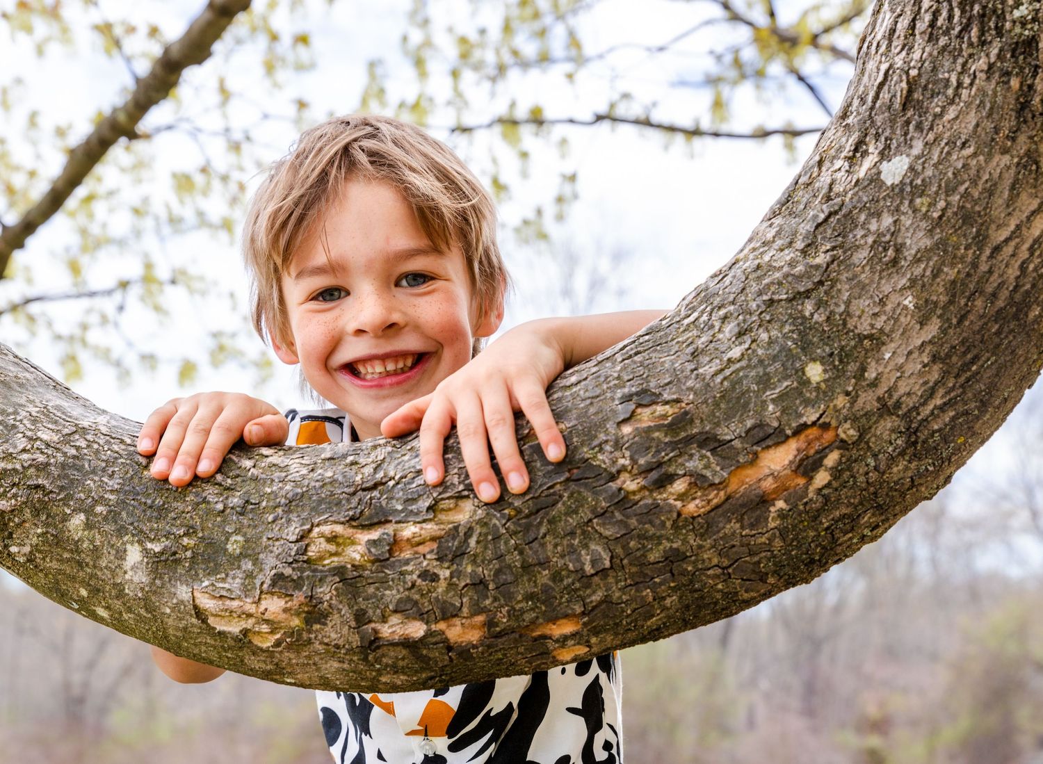 Young person smiling while hanging from thick tree branch in park.