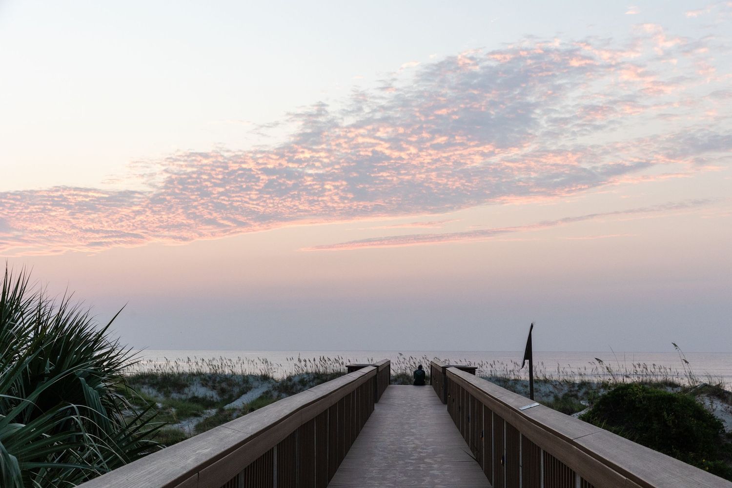 Wooden boardwalk path leads to beach at sunrise with pink clouds and sea grass in Florida.