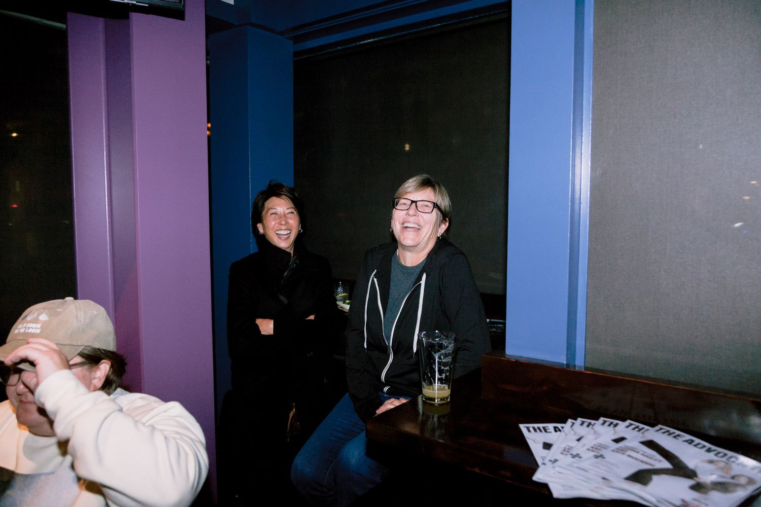 Two people laugh together in a purple-walled venue during a casual indoor social event.