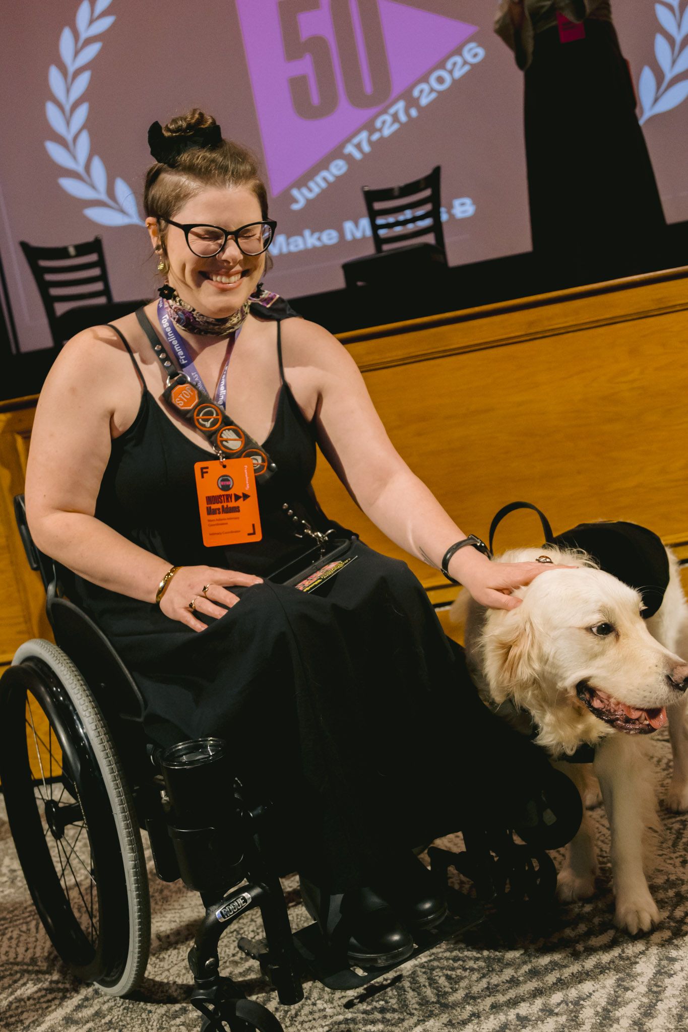 Smiling woman in wheelchair with service dog wearing hat at Frameline50 queer film festival event.