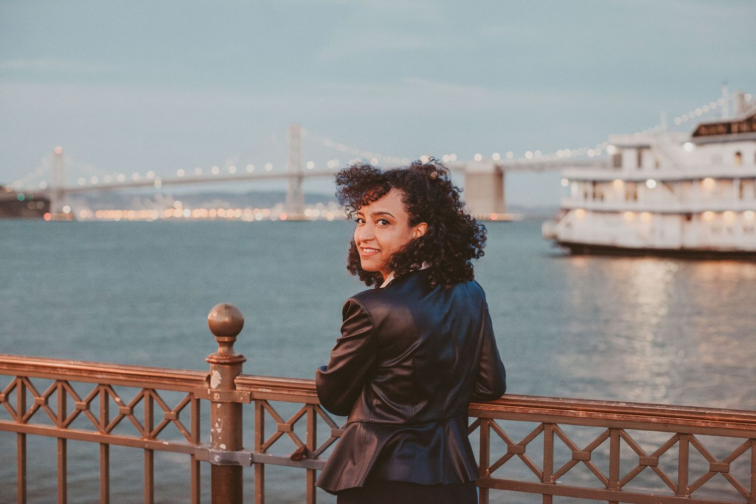 Woman in black leather jacket smiling by waterfront railing with bridge and cruise ship in background.