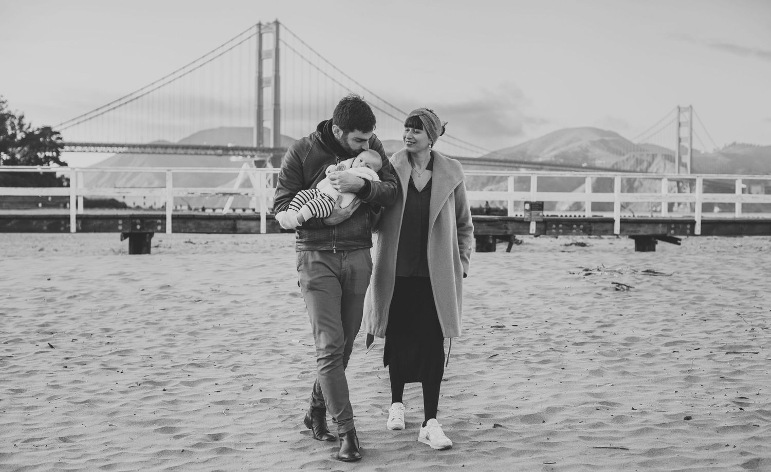 Black and white photo of a couple with newborn baby on a sandy beach with Golden Gate Bridge in background.