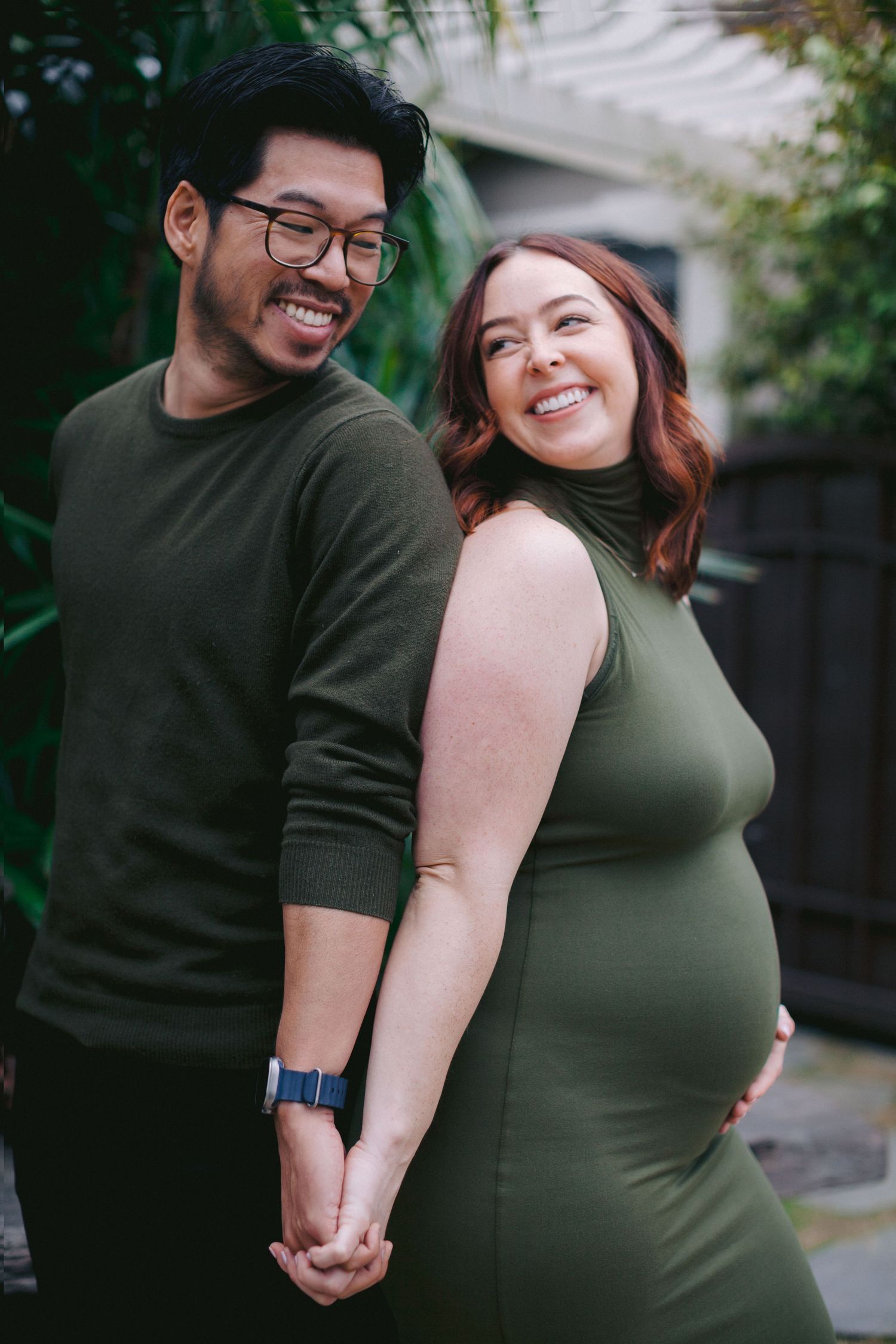 Smiling pregnant couple in matching olive green outfits holding hands outdoors near lush greenery.