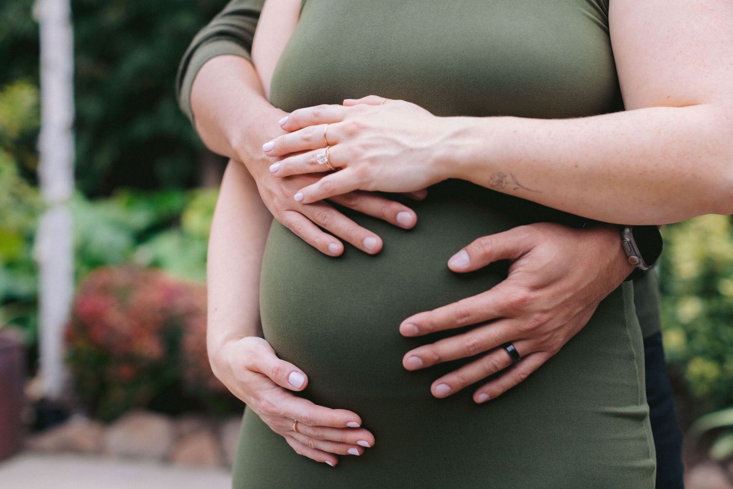 Pregnant woman in green dress cradling baby bump with both hands outdoors in garden setting.