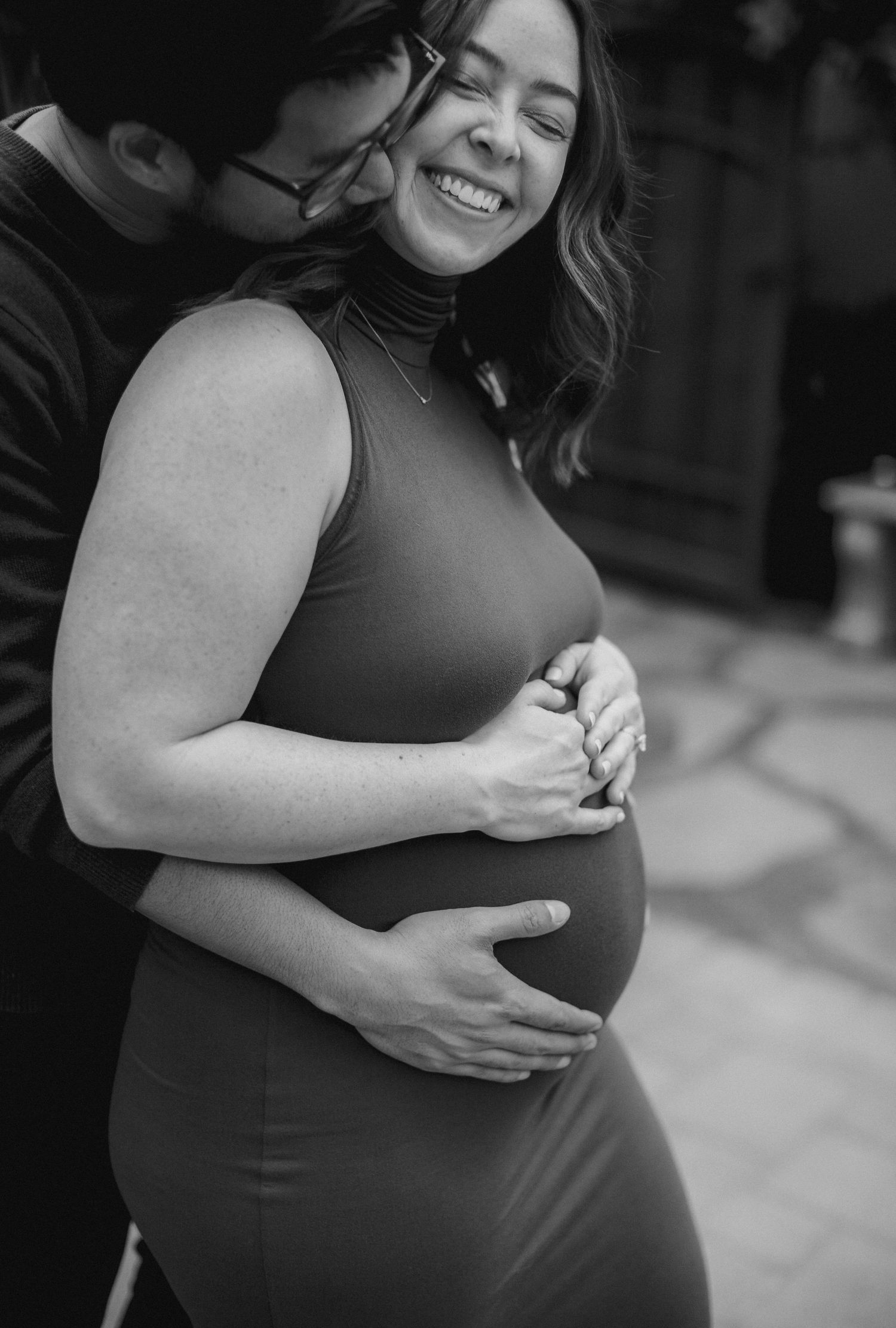 Black and white maternity photo of smiling pregnant woman in fitted dress, embraced by partner cradling her baby bump.