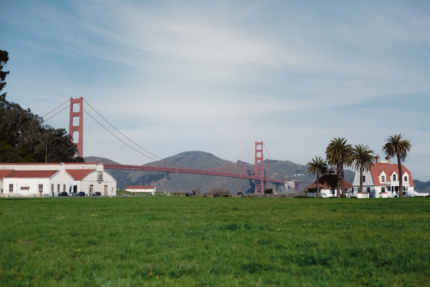 Golden Gate Bridge viewed from Crissy Field, San Francisco, with green lawn, palm trees, and historic white buildings.