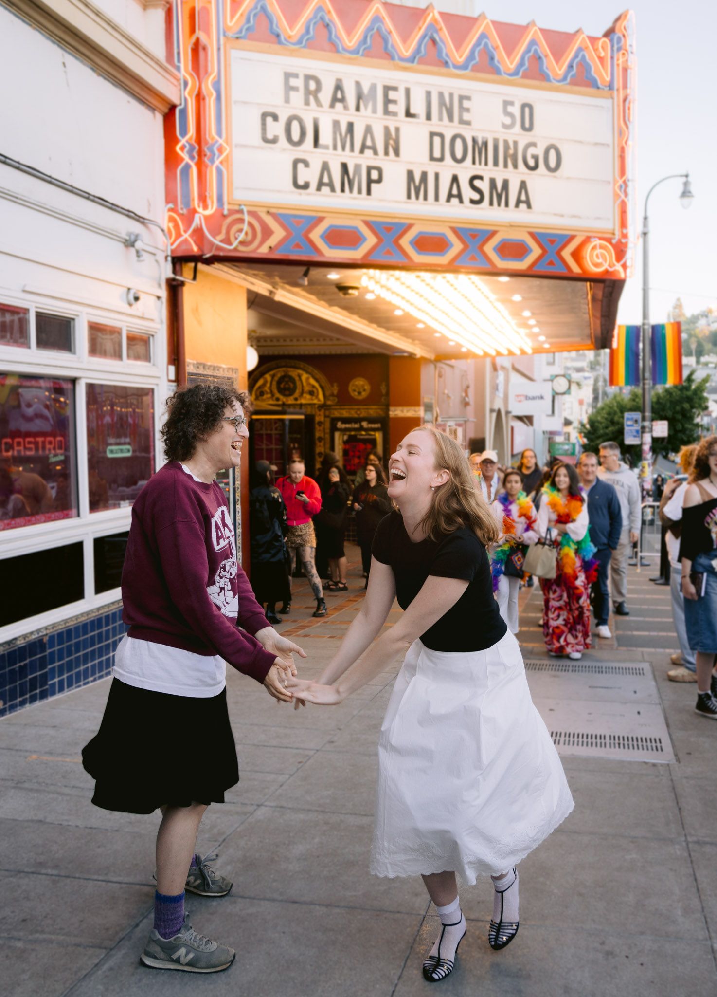 Two people laugh and hold hands outside the Castro Theatre displaying the Frameline 50 marquee for the film Camp Miasma.