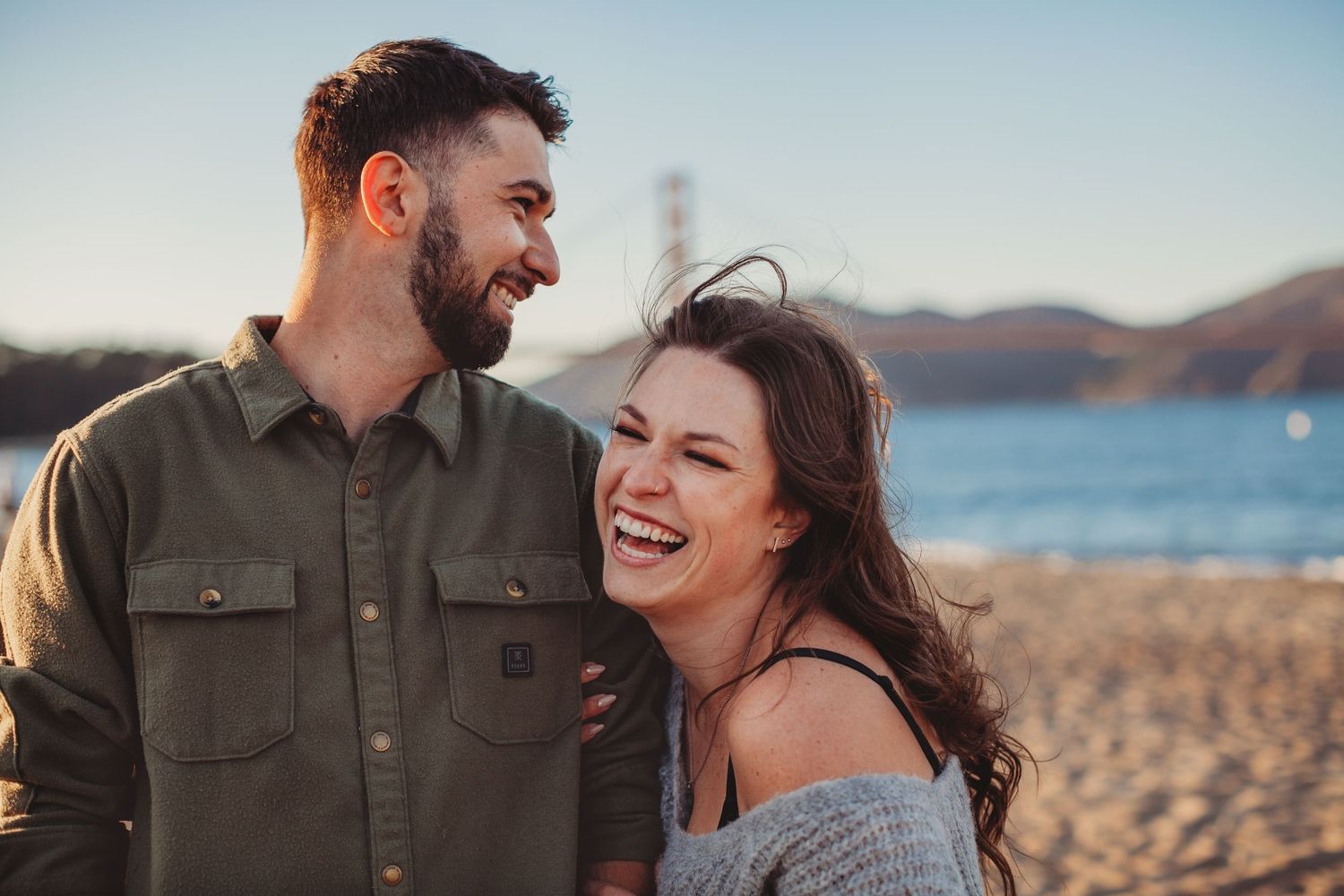 Happy couple laughing on a beach with the Golden Gate Bridge visible in the background.