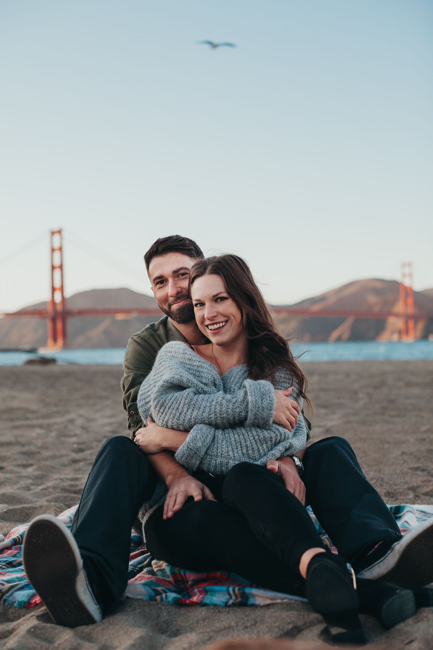 Couple sitting on beach blanket embracing with Golden Gate Bridge in background.