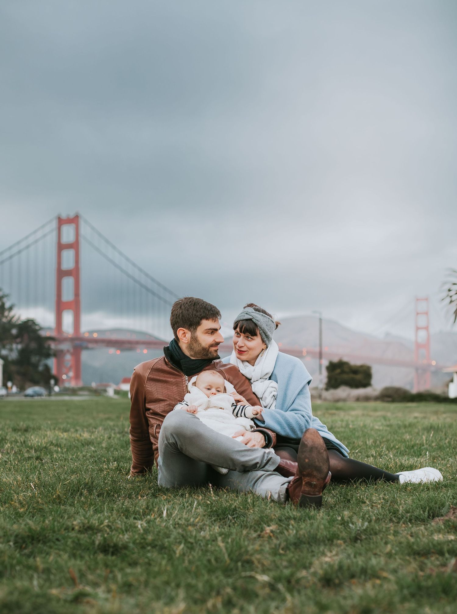 Young family with newborn baby sitting on grass with the Golden Gate Bridge in background.