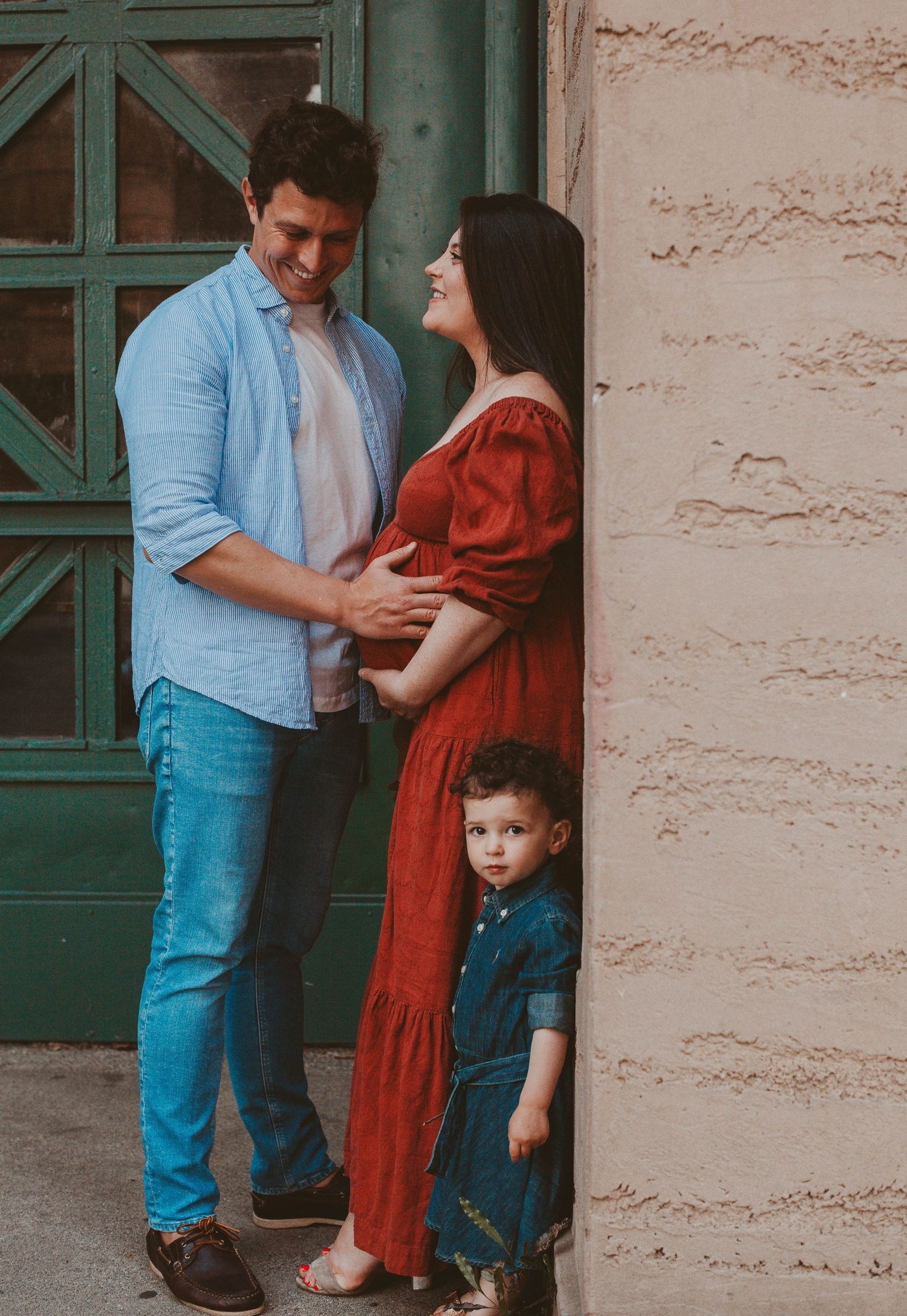 Pregnant woman in red dress with partner and toddler son posing for maternity photo session outdoors.