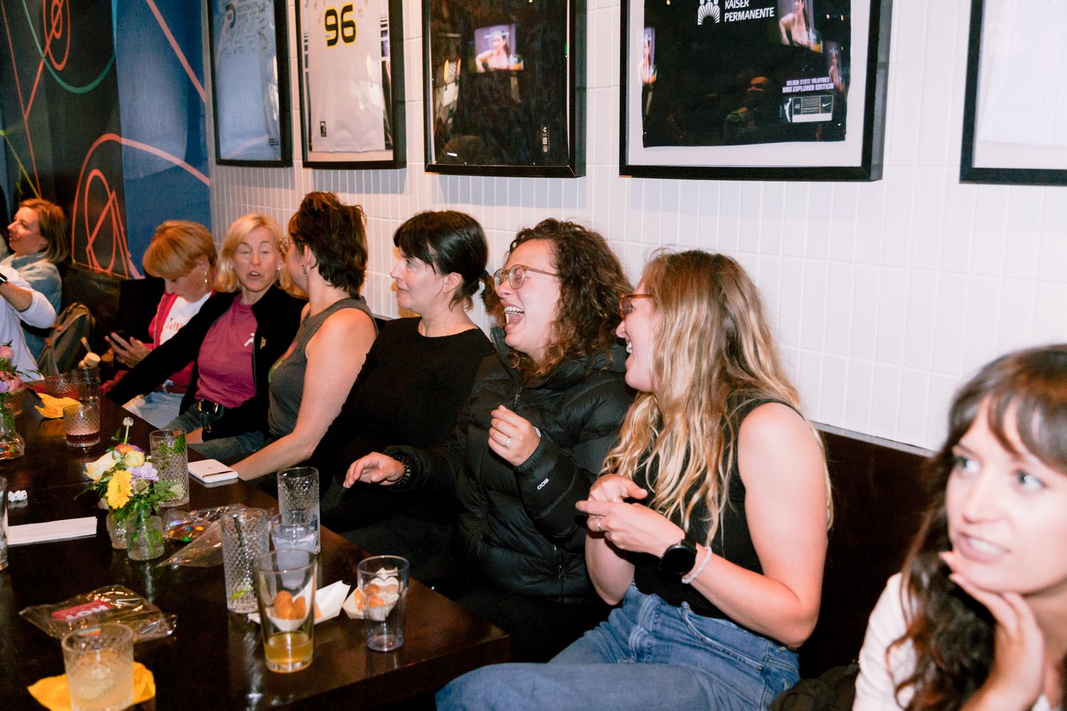 A group of women laugh and socialize together at a bar with drinks on the table.