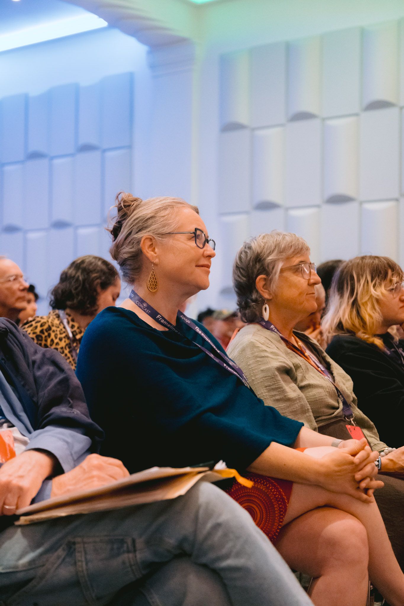 Two older women with gray hair wearing lanyards seated attentively at an indoor conference.
