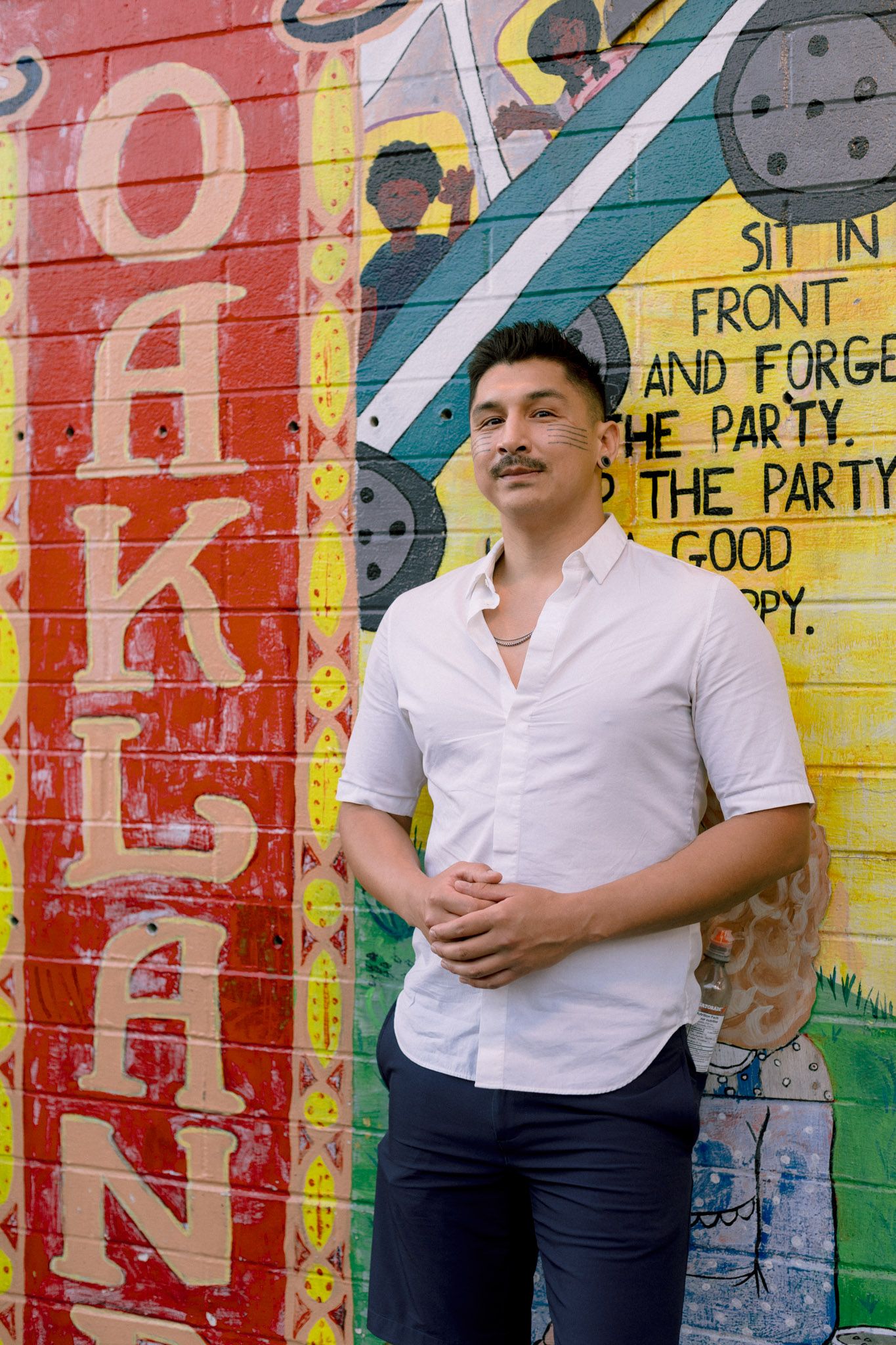 Man with mustache in white shirt posing before colorful Oakland mural with red, yellow, and green colors.