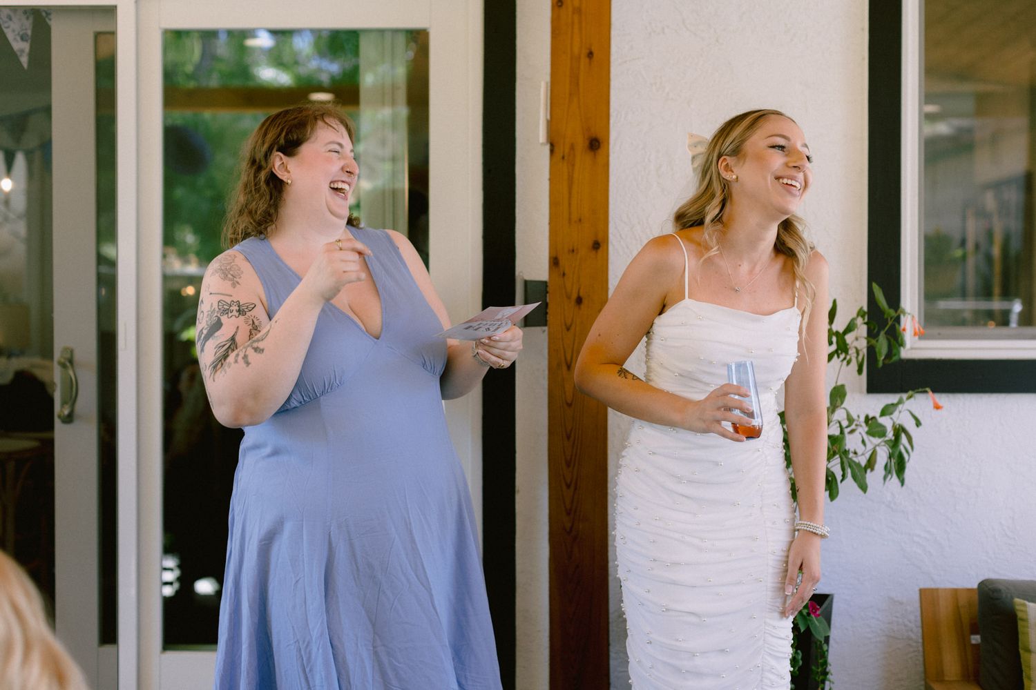 Bride in white dress and bridesmaid in blue laughing joyfully during wedding reception speech indoors.