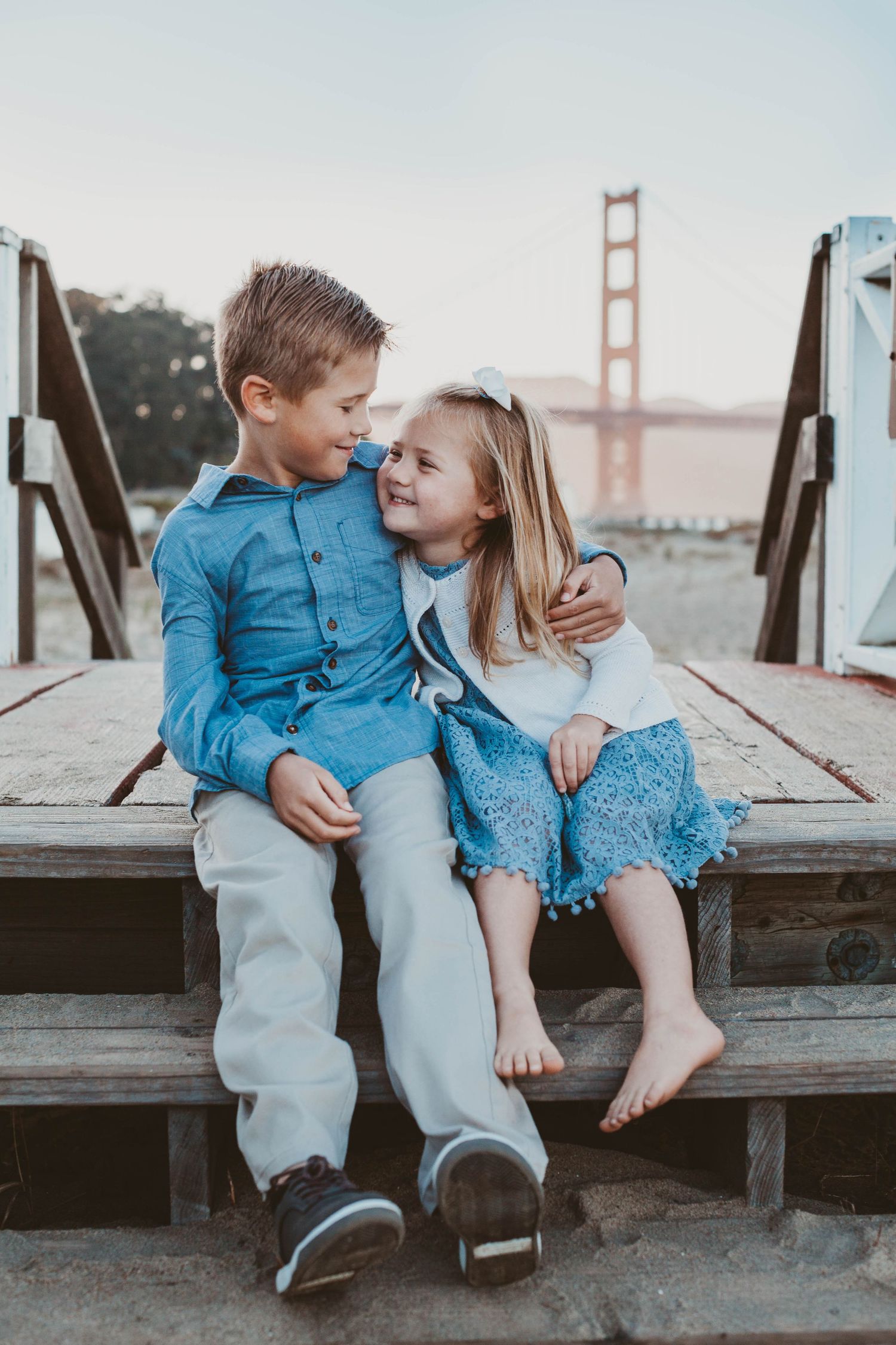 Two young siblings hugging and smiling together on wooden steps with Golden Gate Bridge in background.