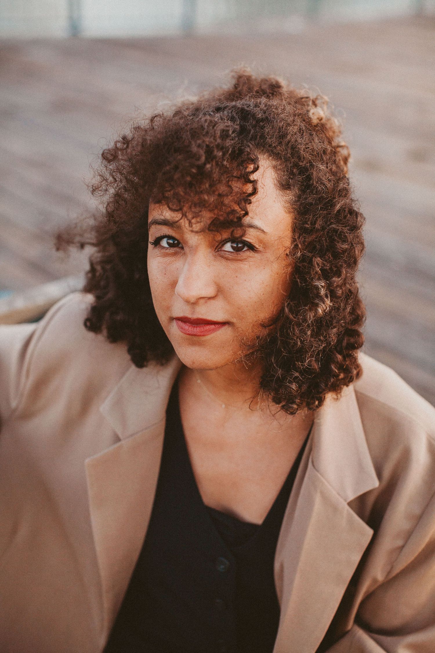 Young woman with curly auburn hair wearing a beige blazer over black top, looking directly at camera outdoors.