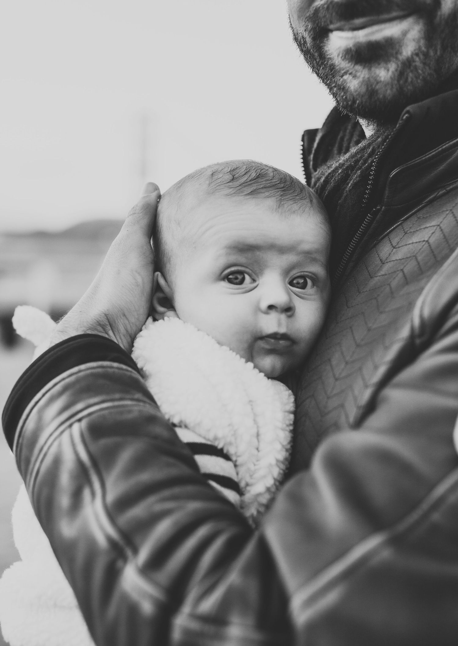 Black and white portrait of a bearded father cradling a newborn baby in a leather jacket.