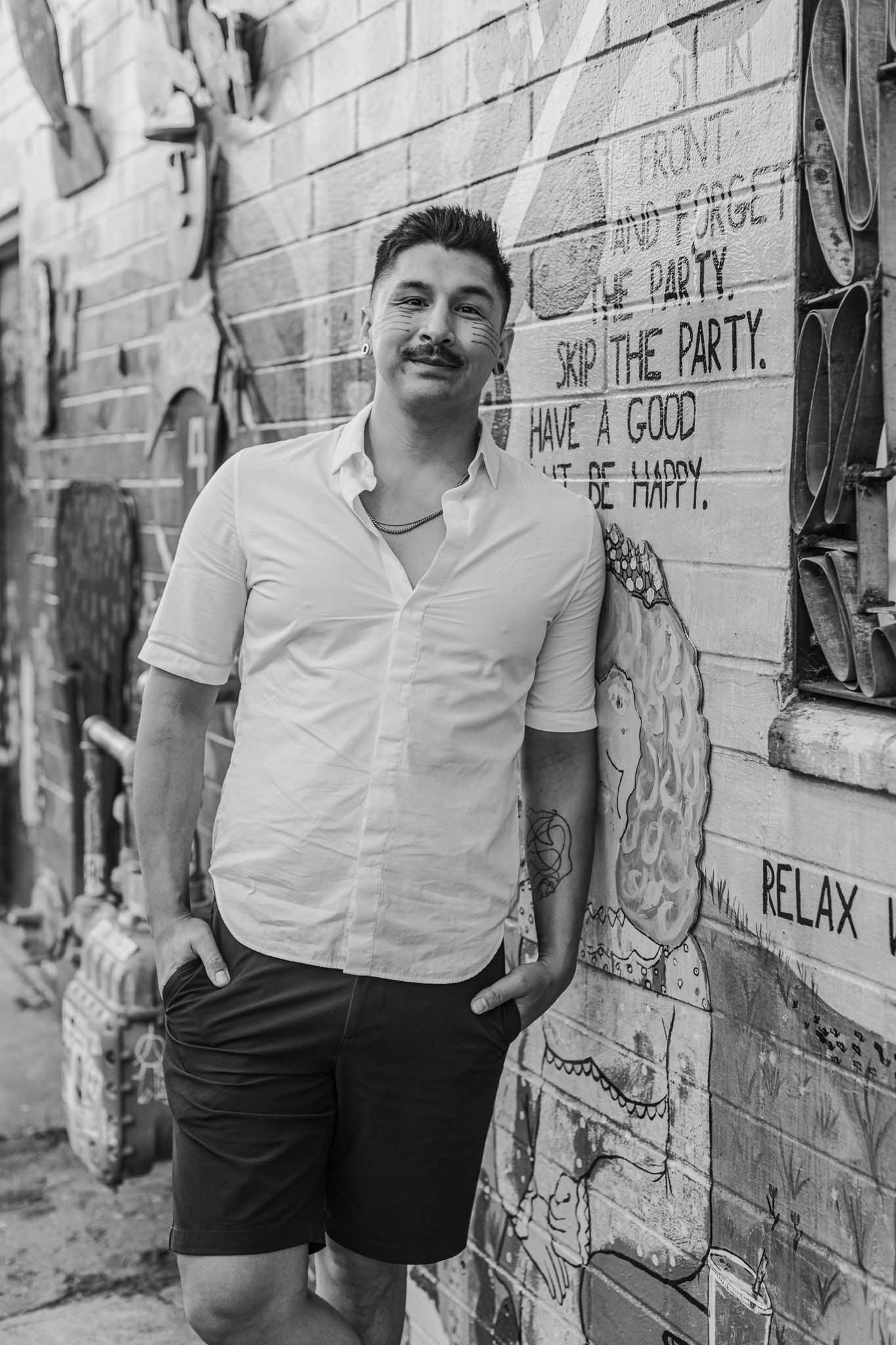 Man with mustache and tattoos leans against graffiti brick wall in black and white photo.