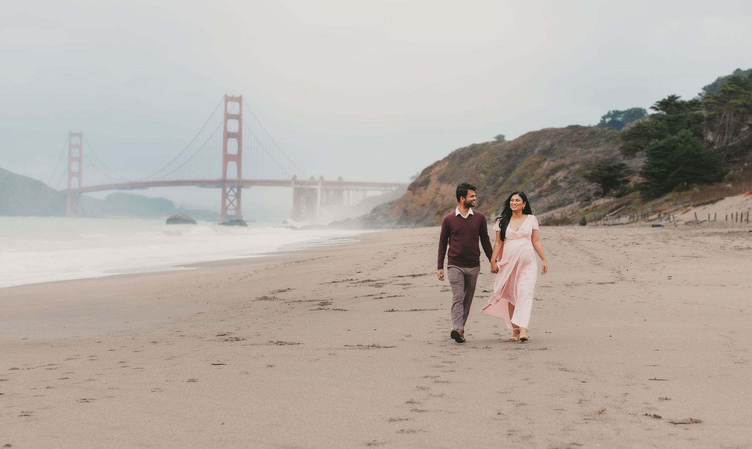 Couple walking hand in hand on Baker Beach with Golden Gate Bridge in misty background, San Francisco.
