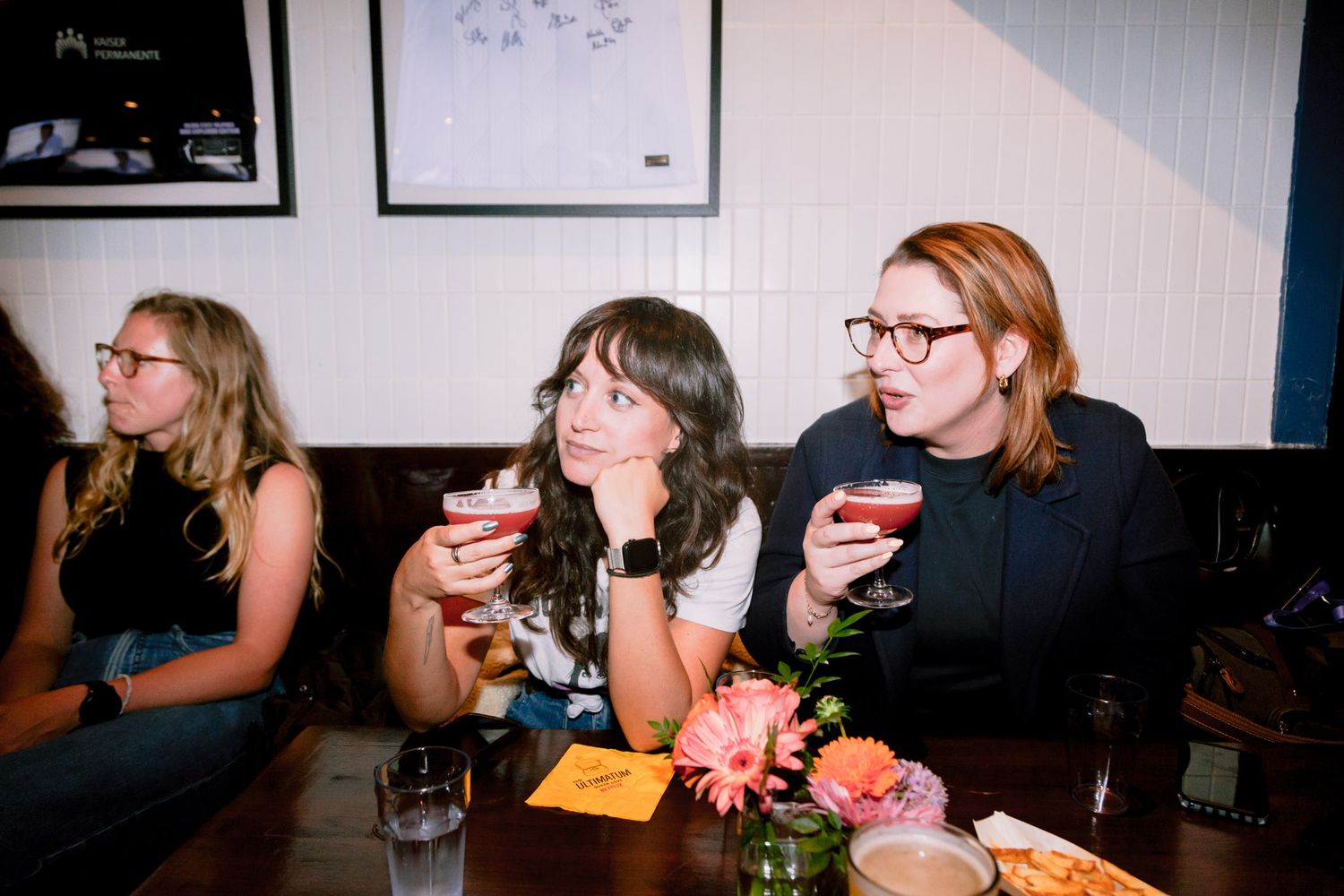 Three women sit at a table holding cocktails, smiling and enjoying a social gathering indoors.