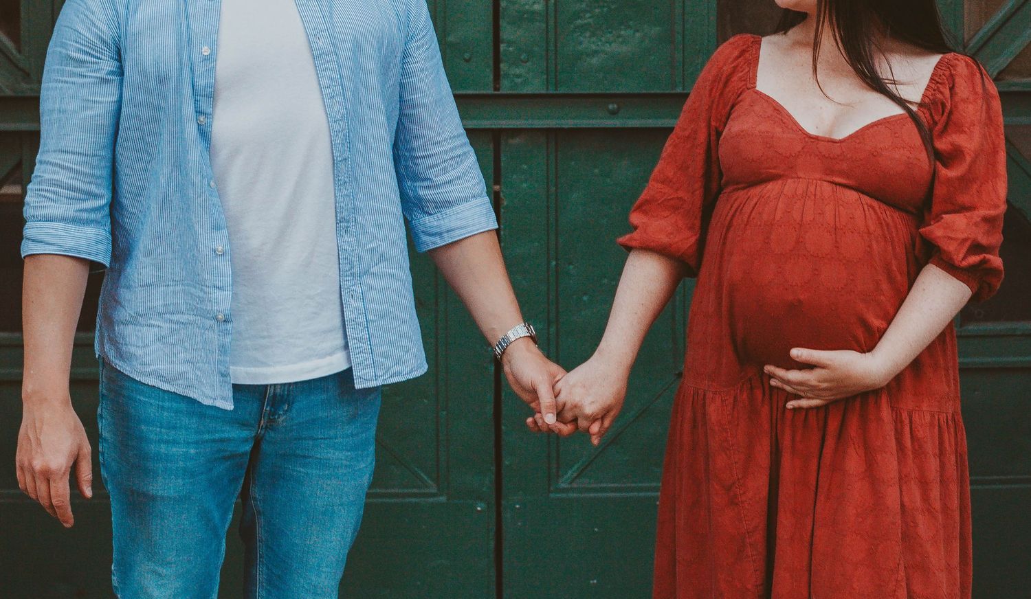 Pregnant woman in red dress holding hands with partner in blue shirt and jeans outdoors.