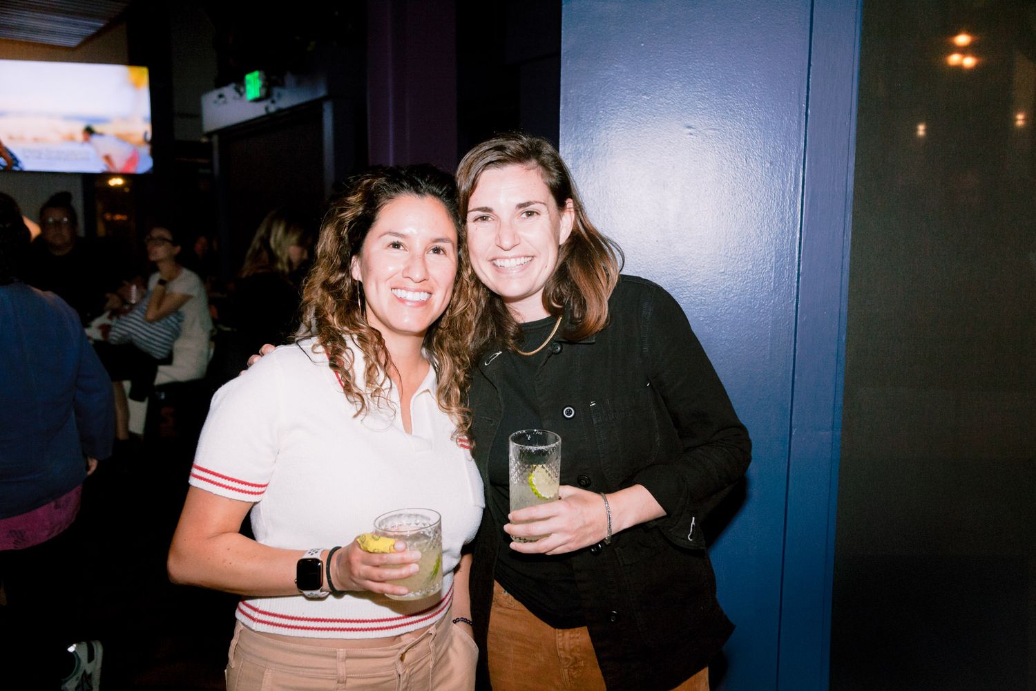 Two women smile and hold cocktails at an indoor social event in a dimly lit venue.