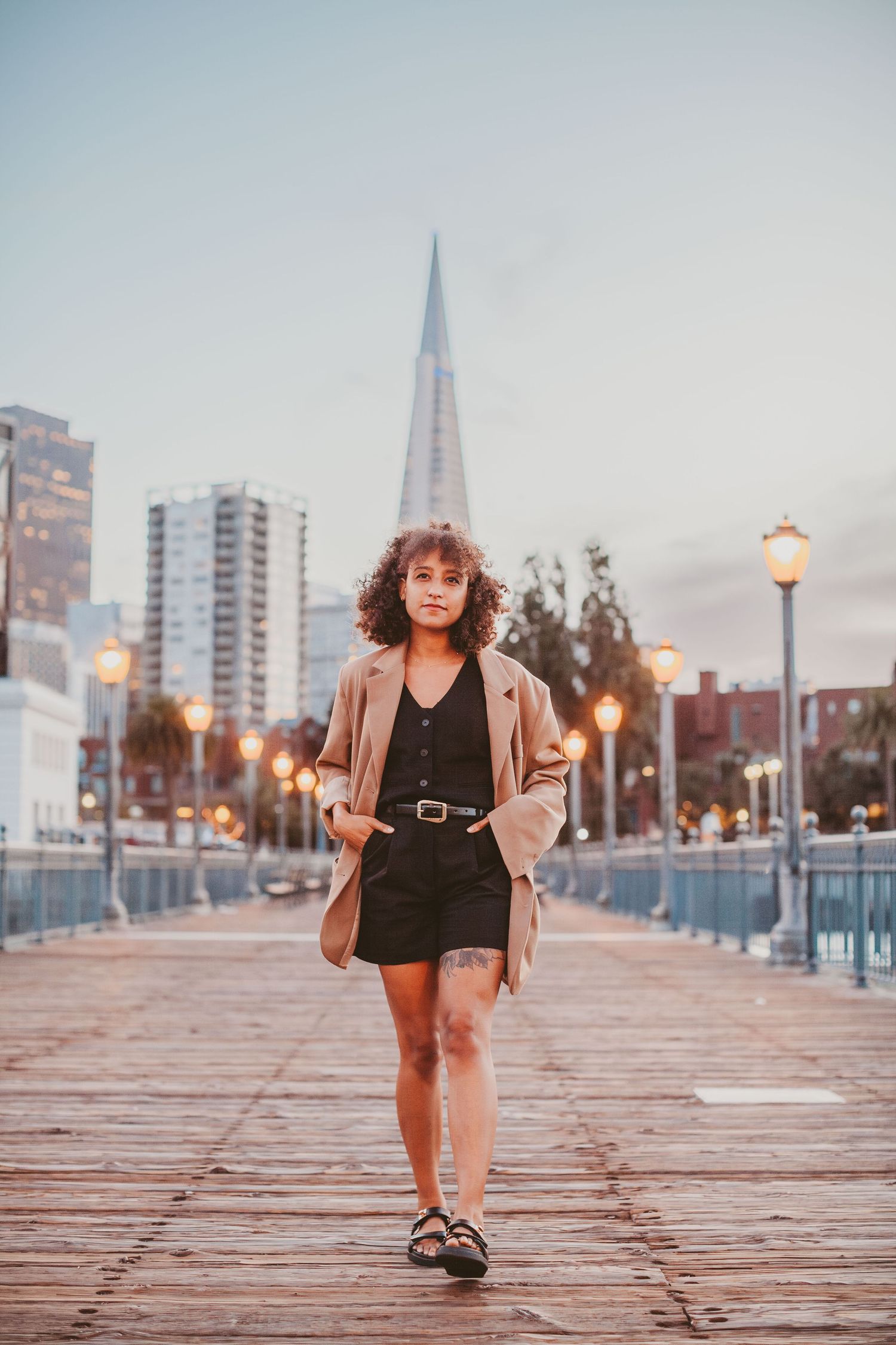 Woman in tan blazer and black shorts walks on waterfront pier with city skyline and Transamerica Pyramid at dusk.