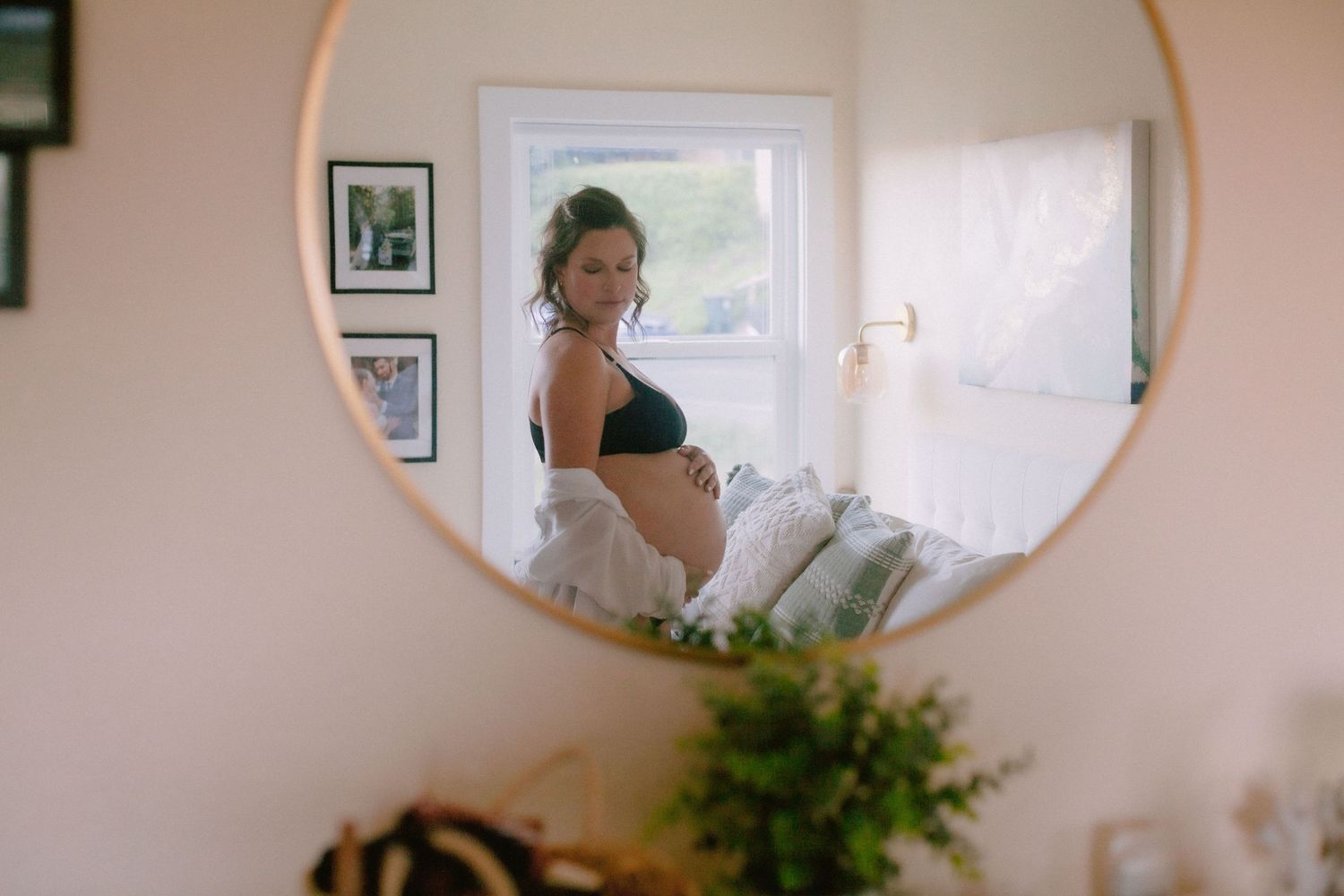 Pregnant woman in black bra reflected in round gold mirror showing baby bump.