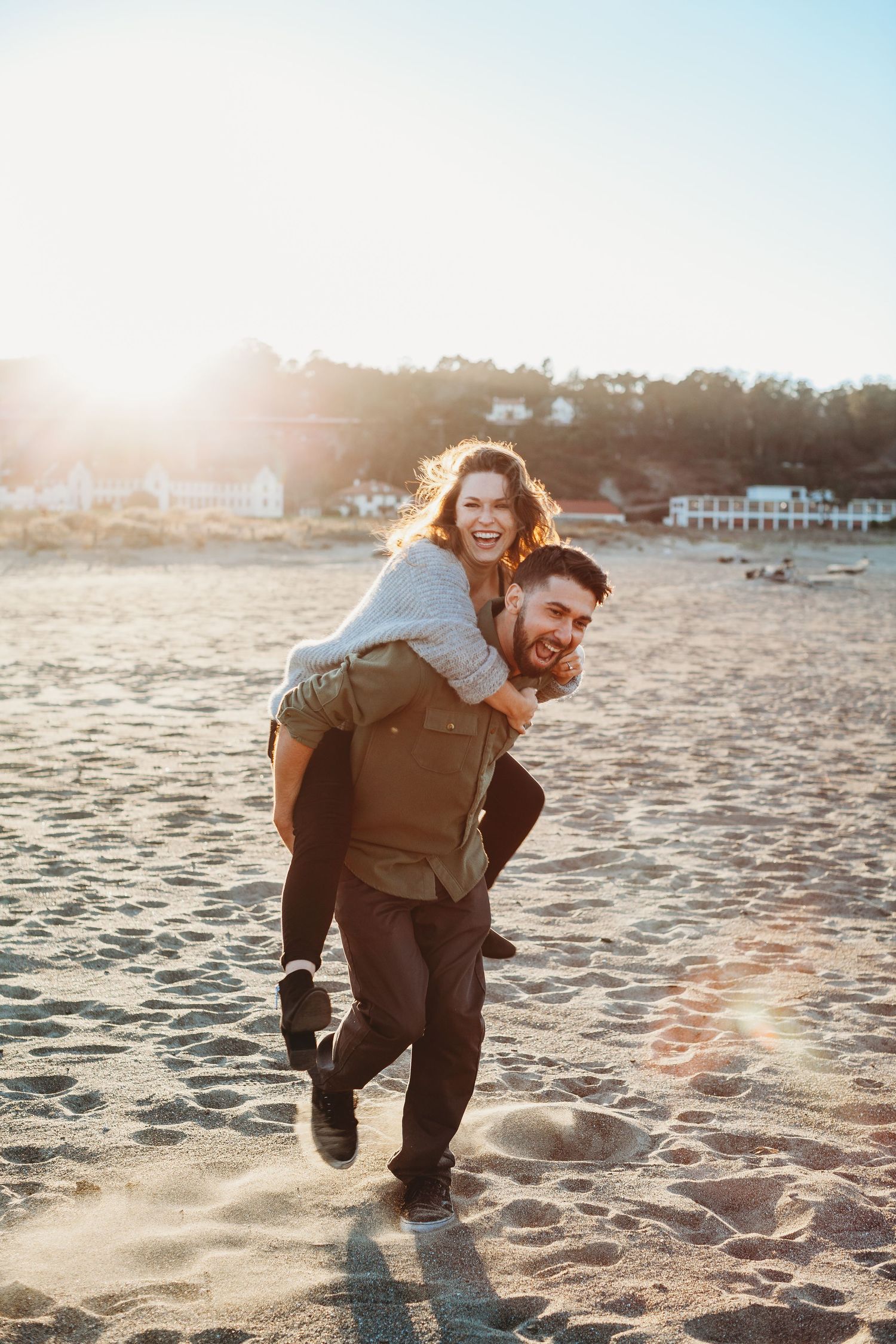 Man giving woman piggyback ride on sunny beach with lens flare.