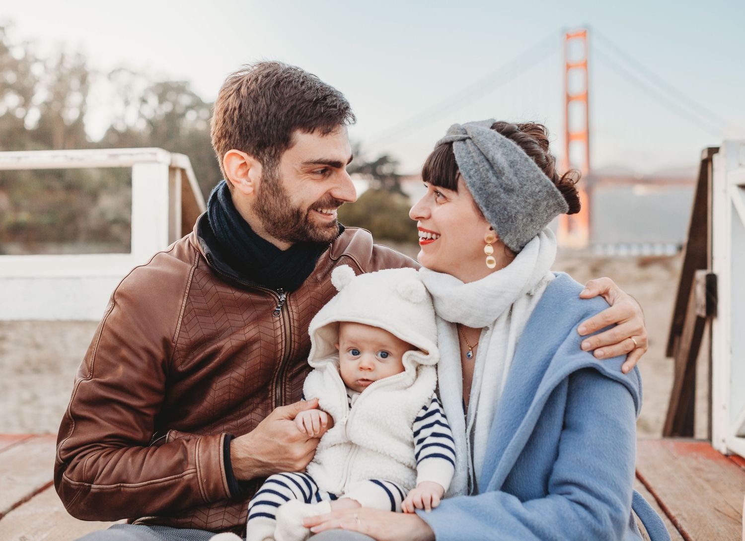 Happy couple sitting on wooden deck with infant baby and Golden Gate Bridge visible in background.