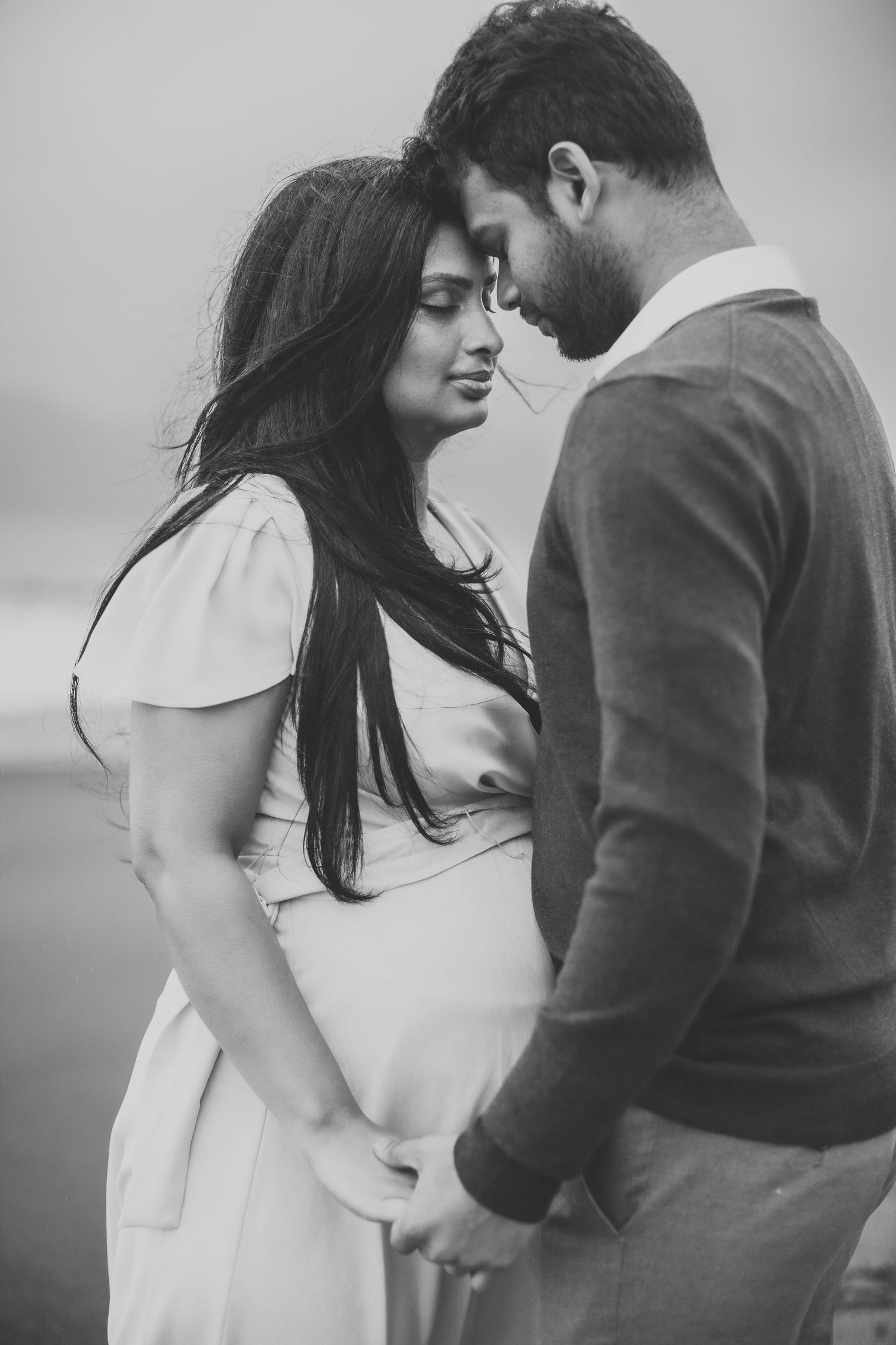 Black and white maternity photo of pregnant couple holding hands and touching foreheads outdoors.