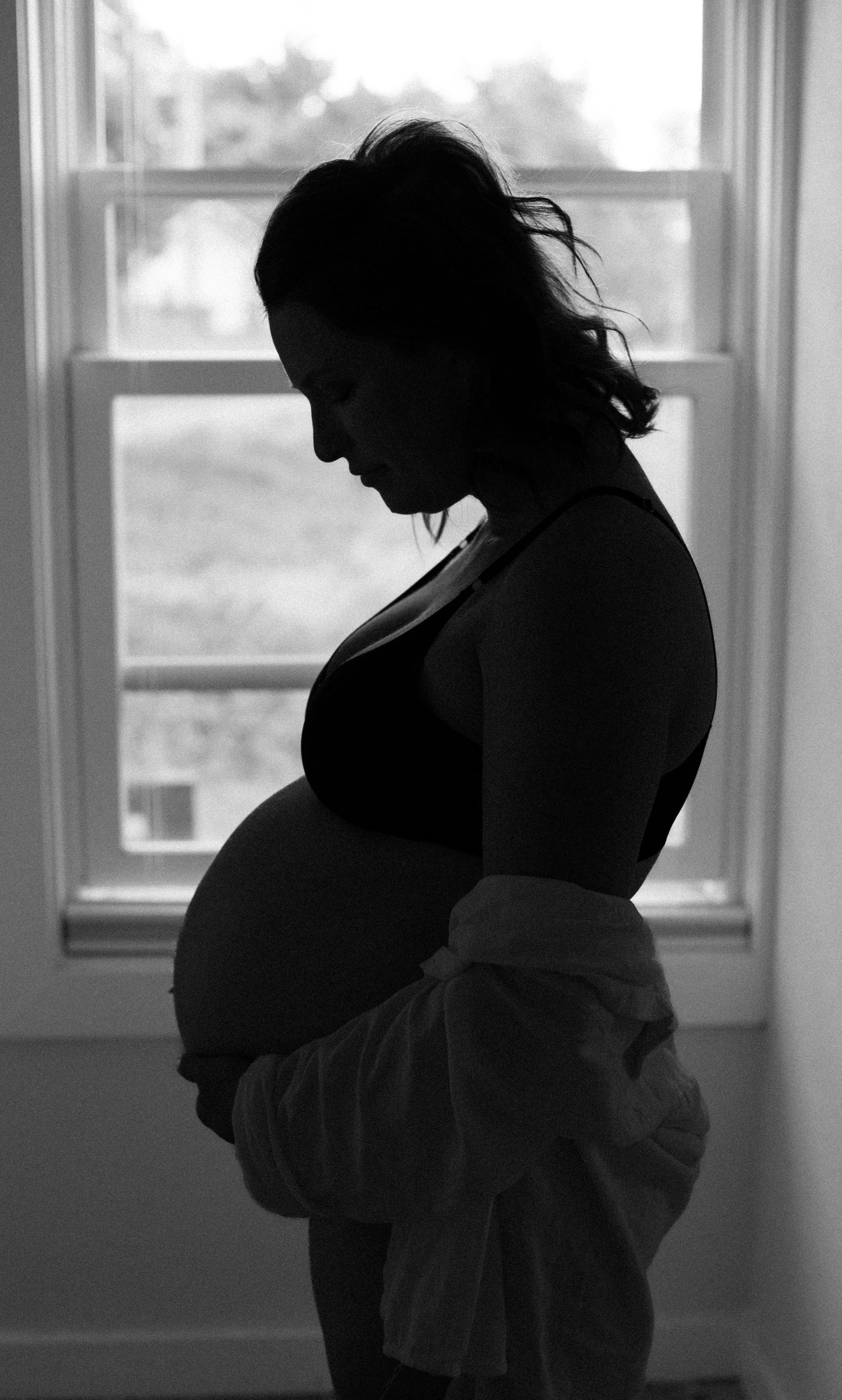 Silhouette of pregnant woman holding belly standing by bright window in black and white.