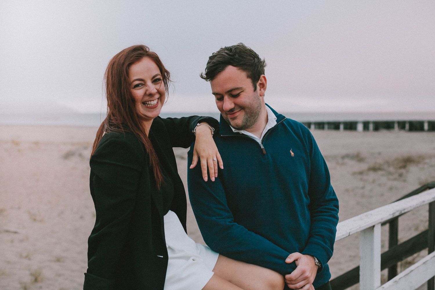 A smiling couple relaxing on a beach boardwalk railing at dusk, woman in black blazer and man in teal pullover.