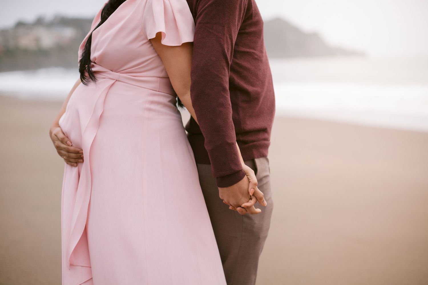 Pregnant couple holding hands on a beach, woman in flowing pink dress with partner in burgundy sweater.