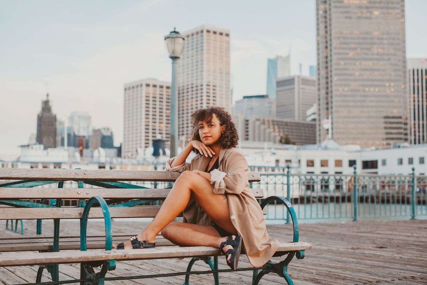 Young woman in trench coat sitting on bench at waterfront pier with city skyline in background.
