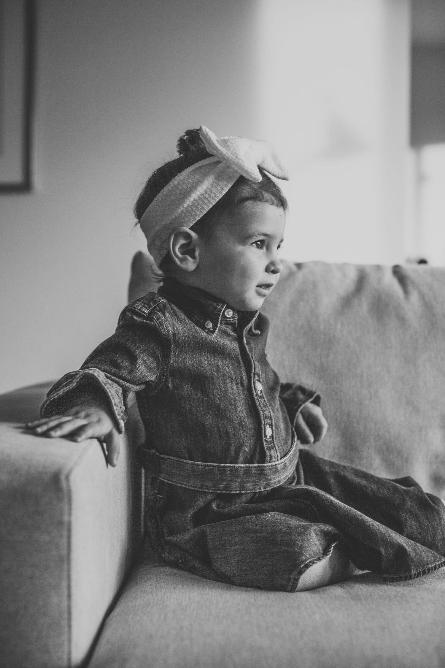 Black and white portrait of a baby in denim outfit and headband sitting on a couch.