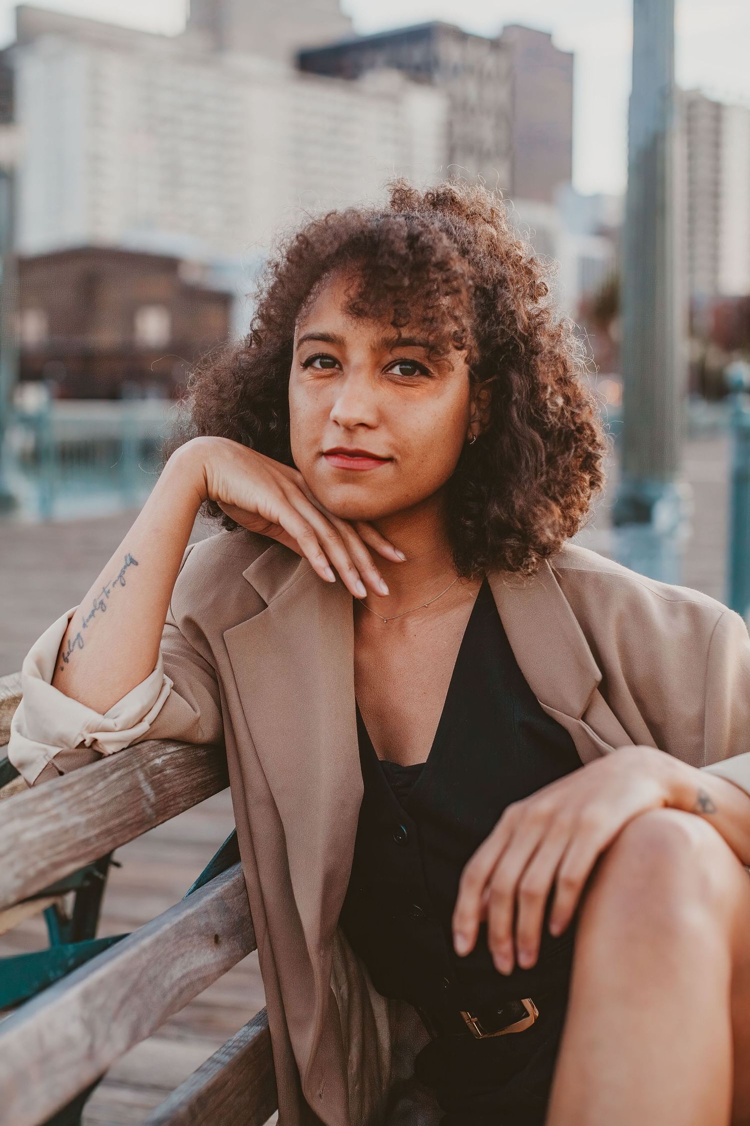 A young woman with curly hair in a beige blazer sits on a bench with a city skyline backdrop.