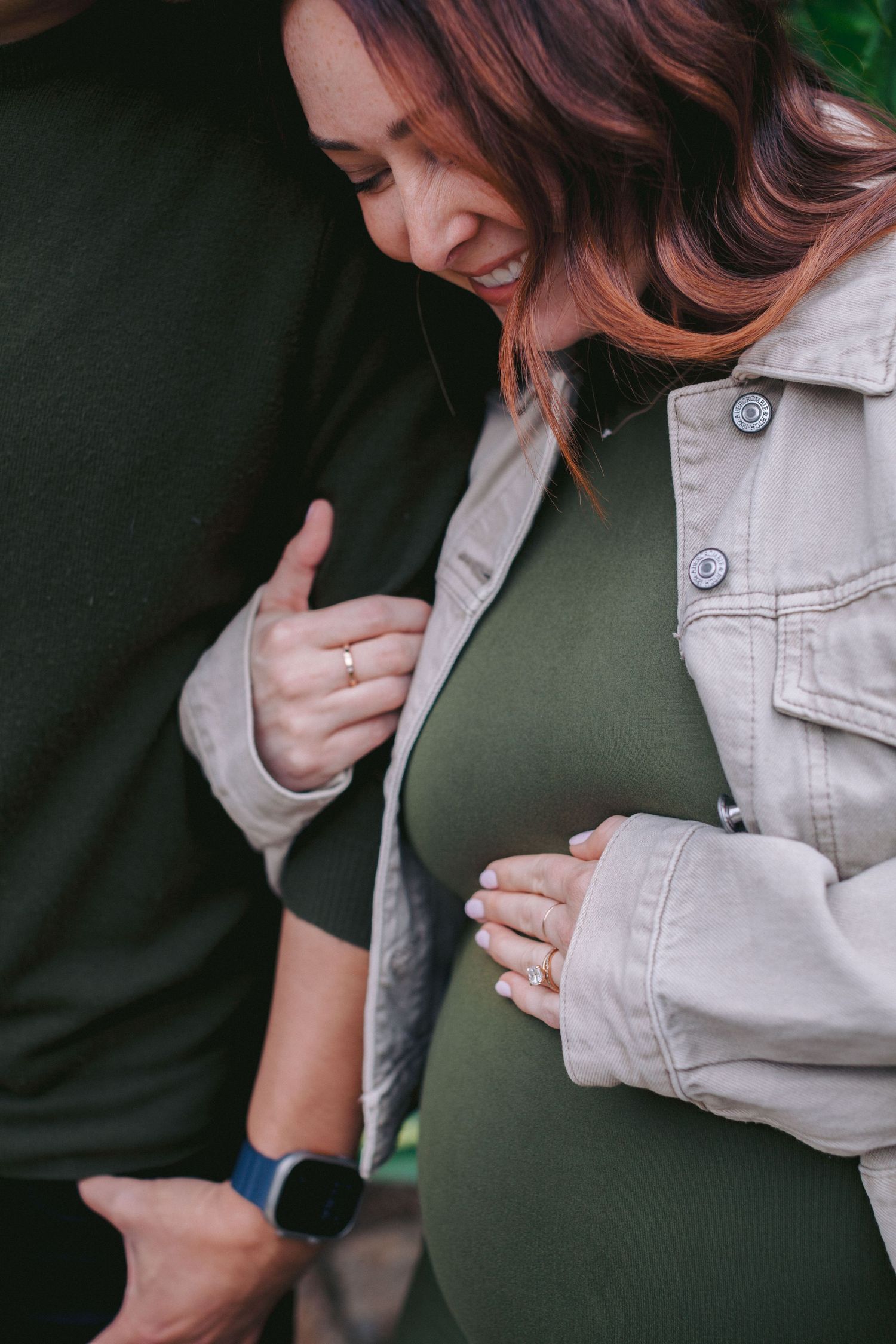 Pregnant woman in olive dress and beige jacket tenderly holds her baby bump while partner embraces her from behind.