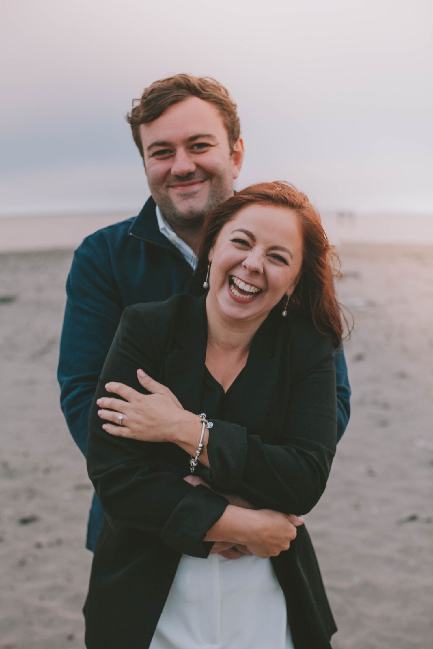 Happy couple embracing on a sandy beach at dusk, woman laughing joyfully while man hugs her from behind.