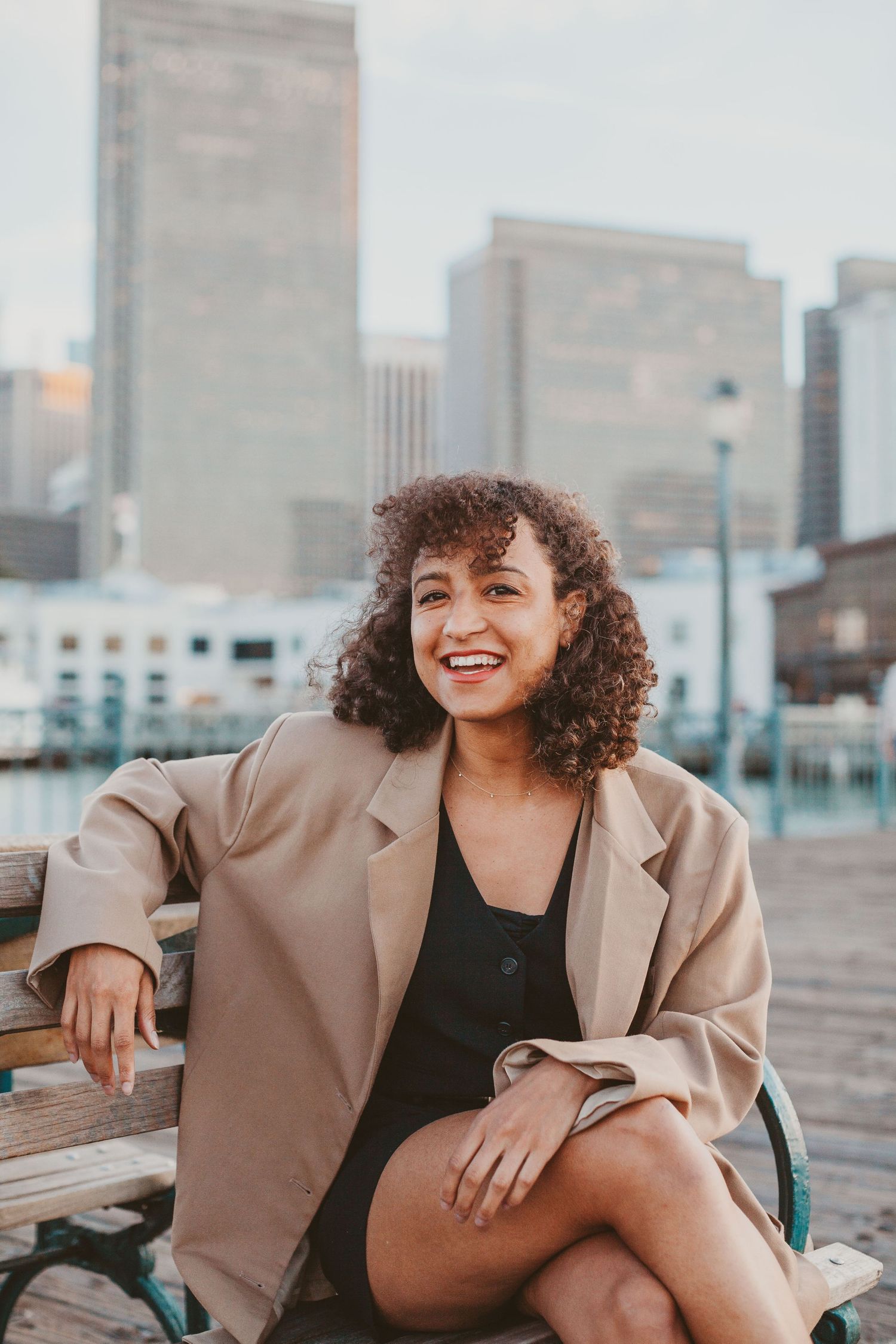 A young woman with curly hair smiles while sitting on a bench with a city skyline in the background.