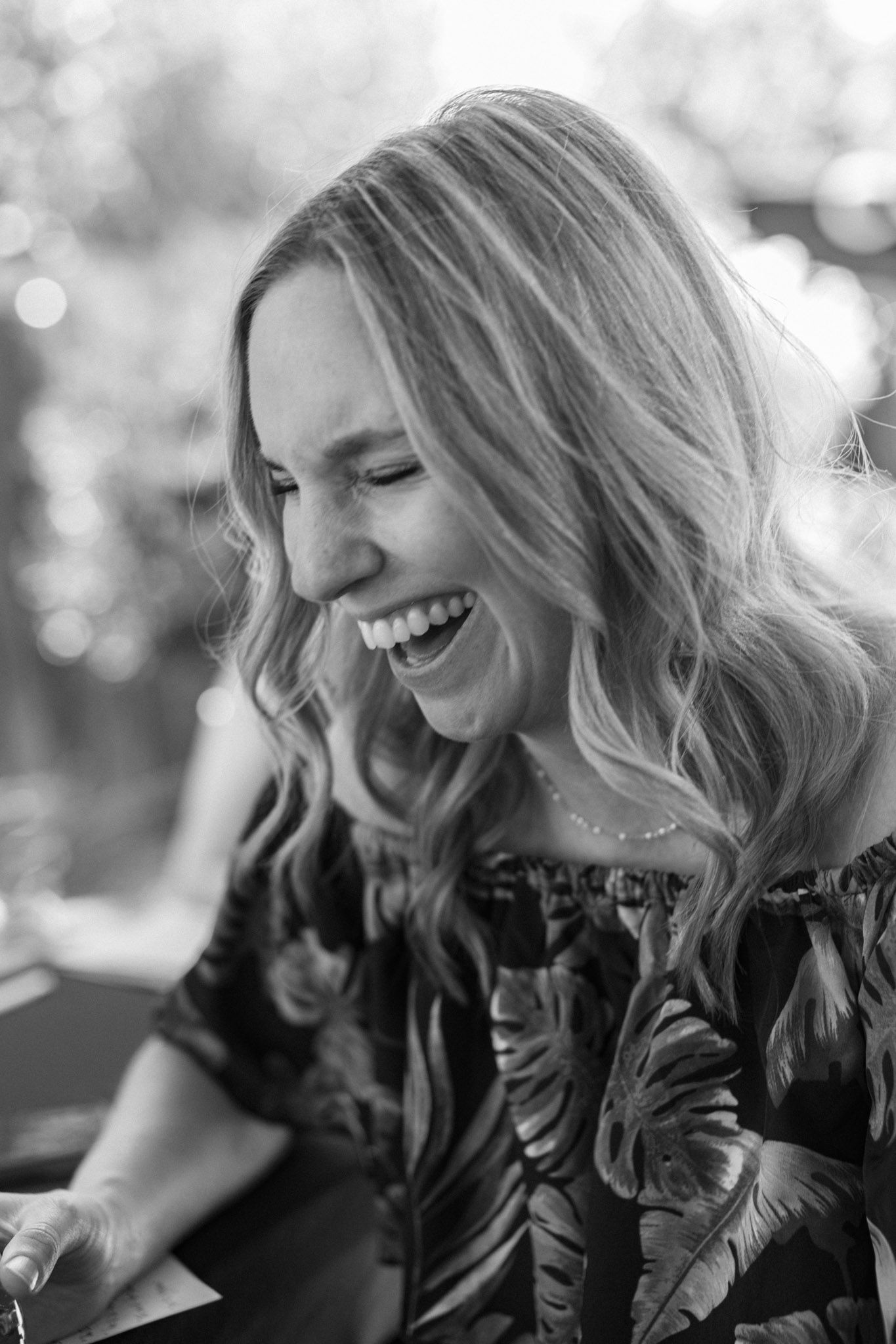 Black and white photo of a laughing blonde woman in a floral off-shoulder top, captured in natural outdoor light.