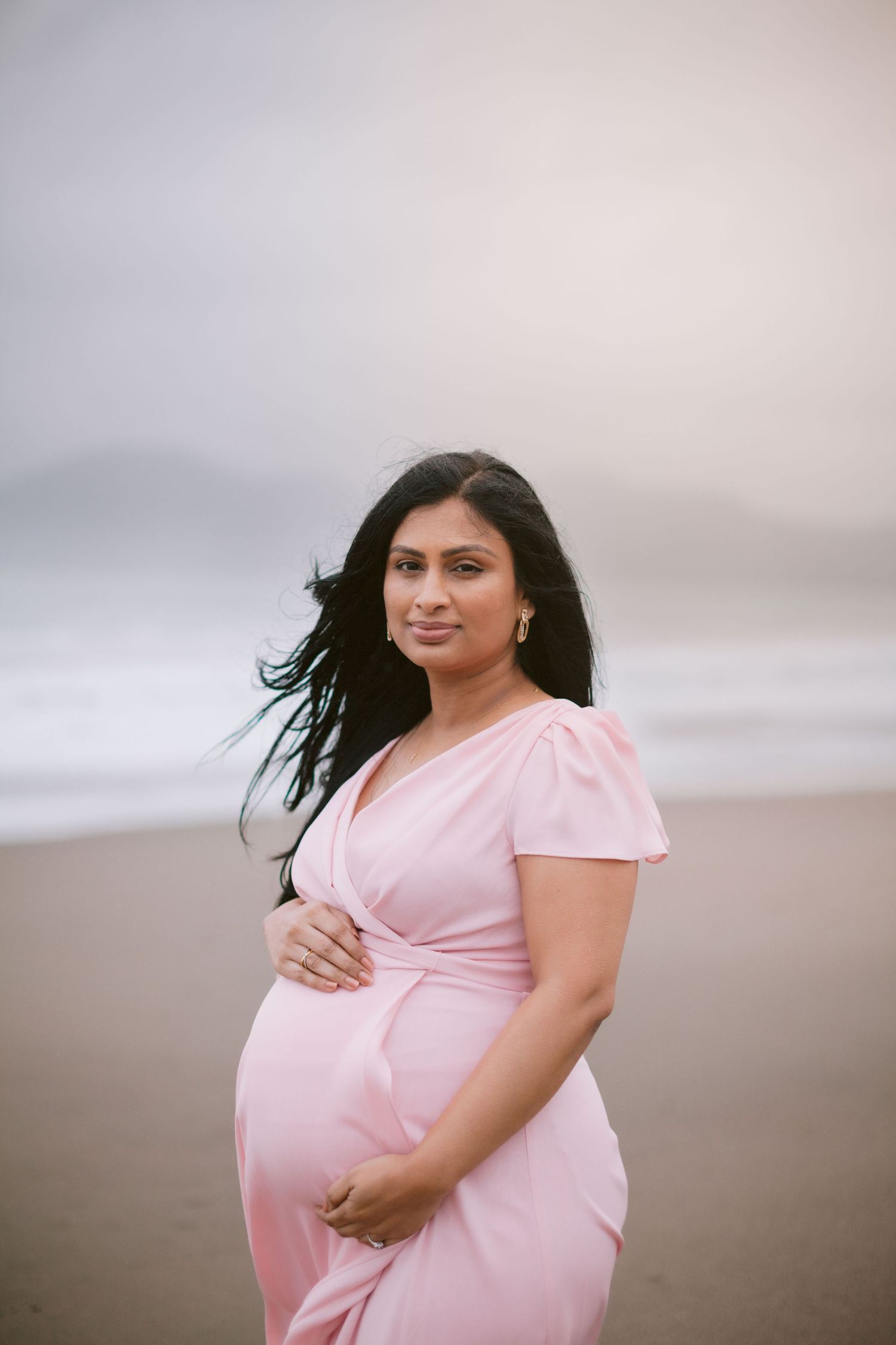 Pregnant woman in pink dress cradling baby bump on misty beach at dusk.