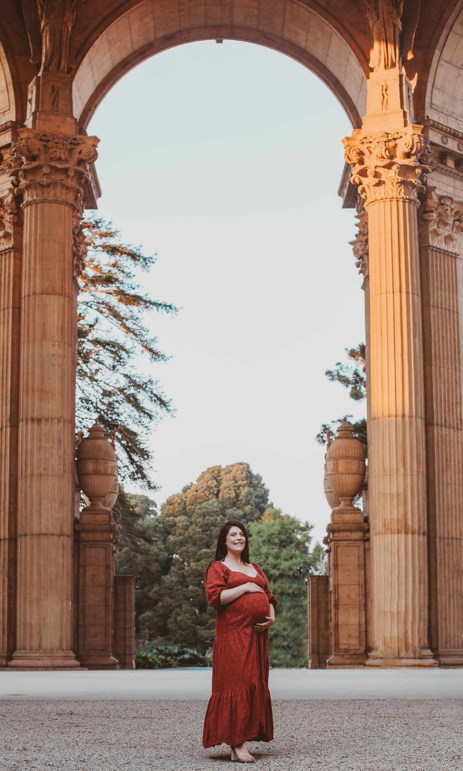 Pregnant woman in red dress posing under ornate stone arch at Palace of Fine Arts, San Francisco.